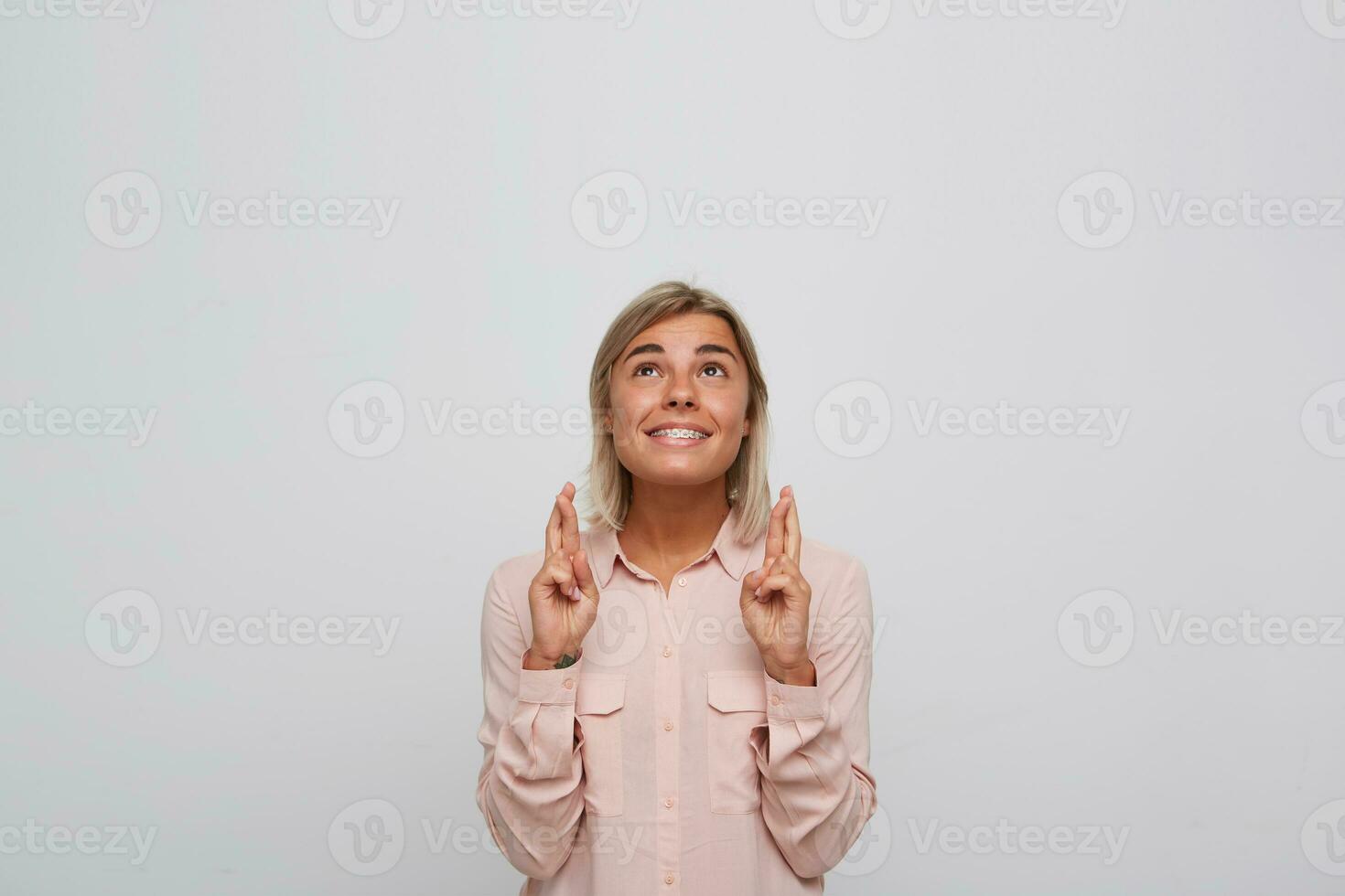 Portrait of happy excited blonde young woman with braces on teeth wears pink shirt keeps fingers crossed, looks up and makes a wish isolated over white background photo