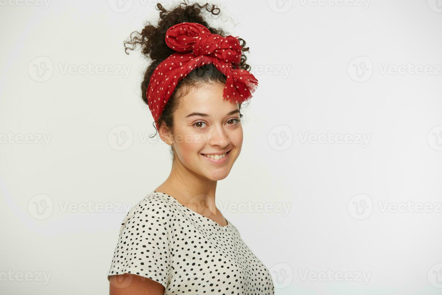 Shot of pretty satisfied young female smiles, rejoices good offer, glad to participate in interesting event, stands sideways, dressed in T-shirt with black polka dots, isolated over white studio wall photo