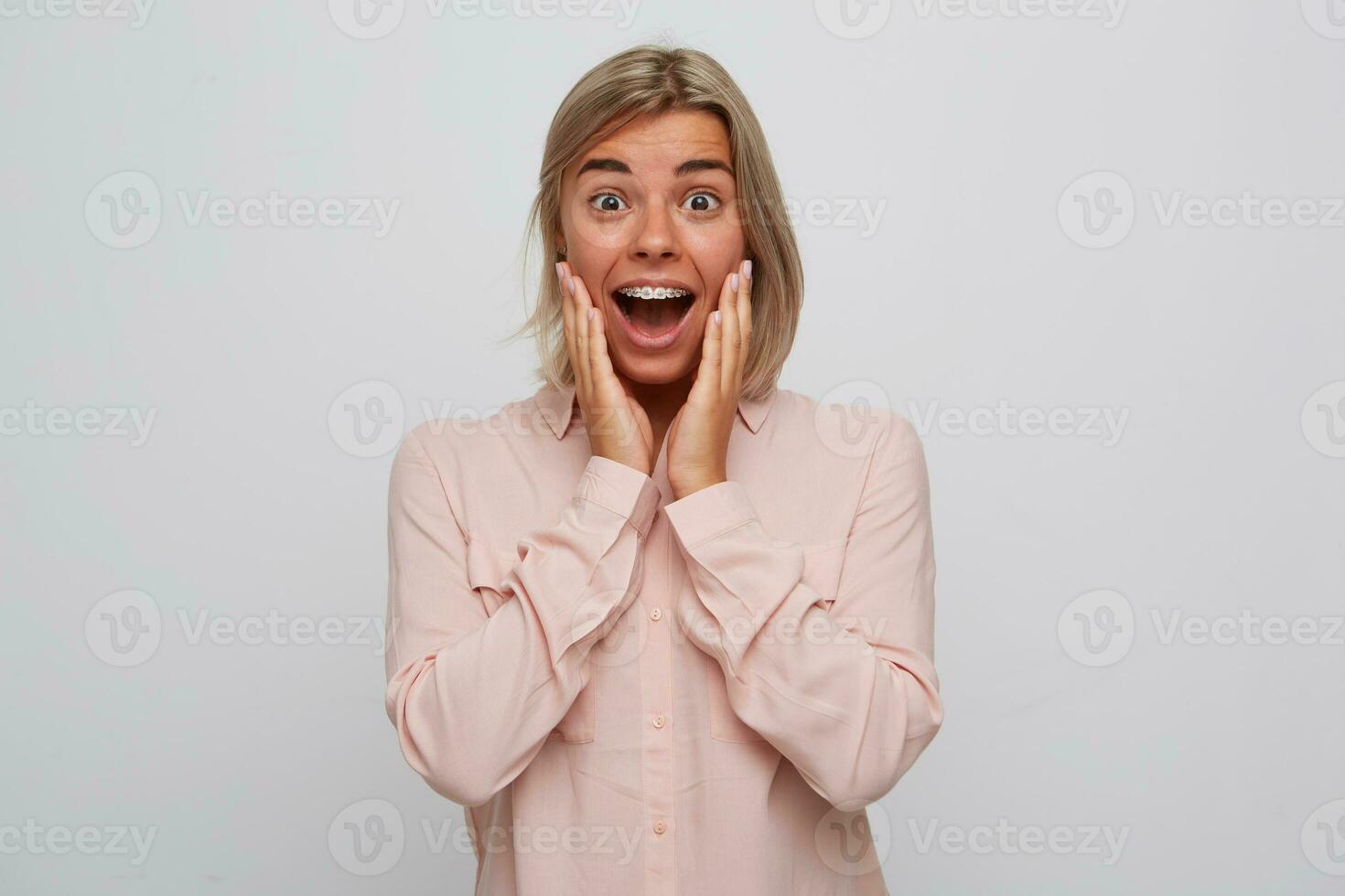 Portrait of cheerful excited blonde young woman with braces on teeth wears pink shirt touching her cheek, looks surprised and shouting isolated over white background photo