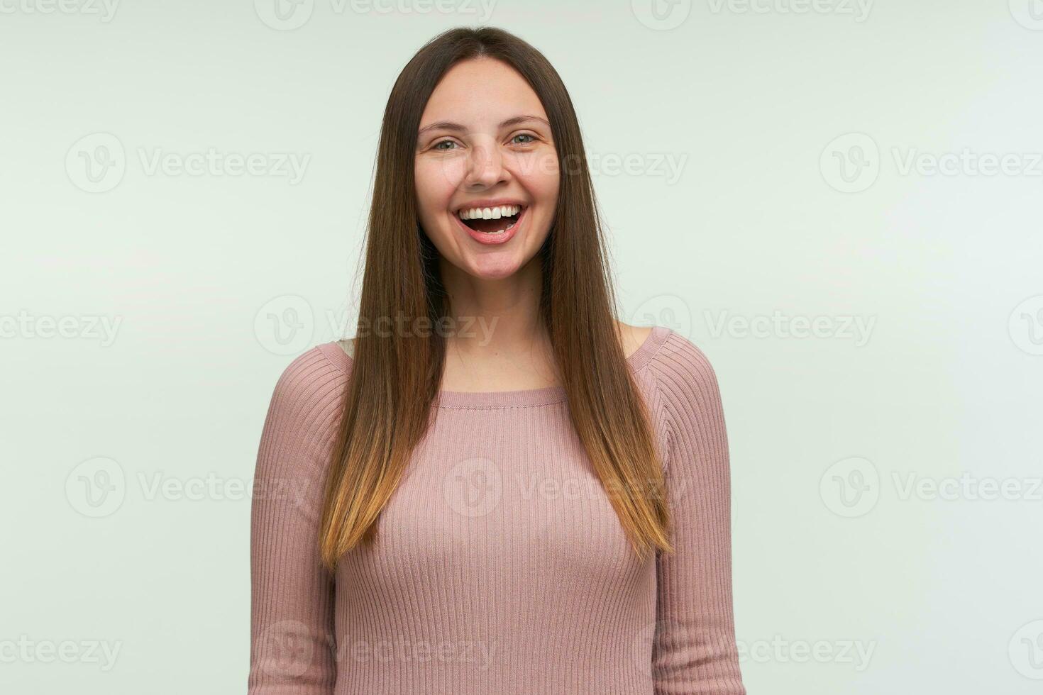 Portrait of young woman with long hair down, looks glad, laughing on a joke, smiling, dressed in a tight pink knit sweater, isolated over white background photo