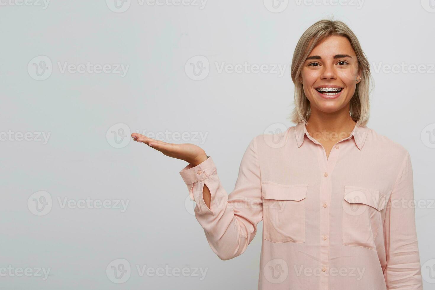 Closeup of happy confident blonde young woman with braces on teeth wears pink shirt smiling and holding copyspace on palm isolated over white background photo