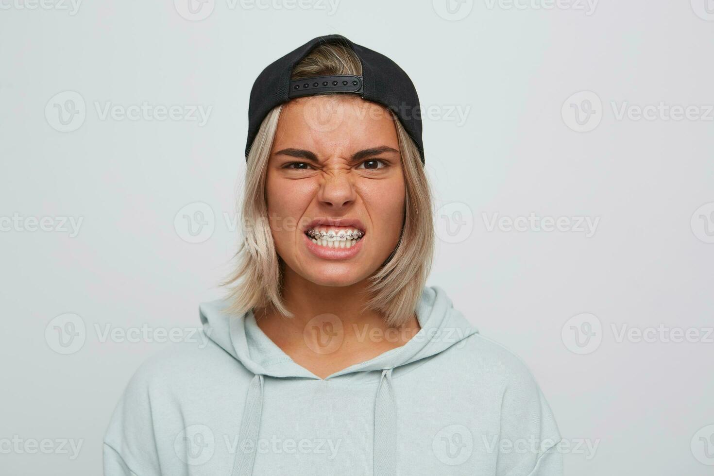 Portrait of mad angry blonde young woman with braces on teeth wears black cap and hoodie looks irritated and furious isolated over white background photo