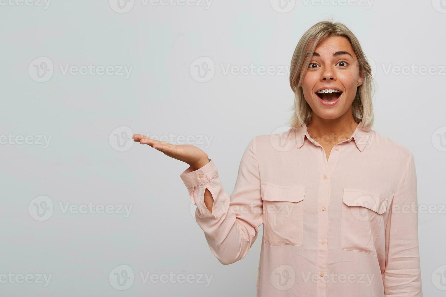 Portrait of smiling amazed blonde young woman with braces on teeth and opened mouth wears pink shirt looks surprised and holding copyspace on palm isolated over white background photo