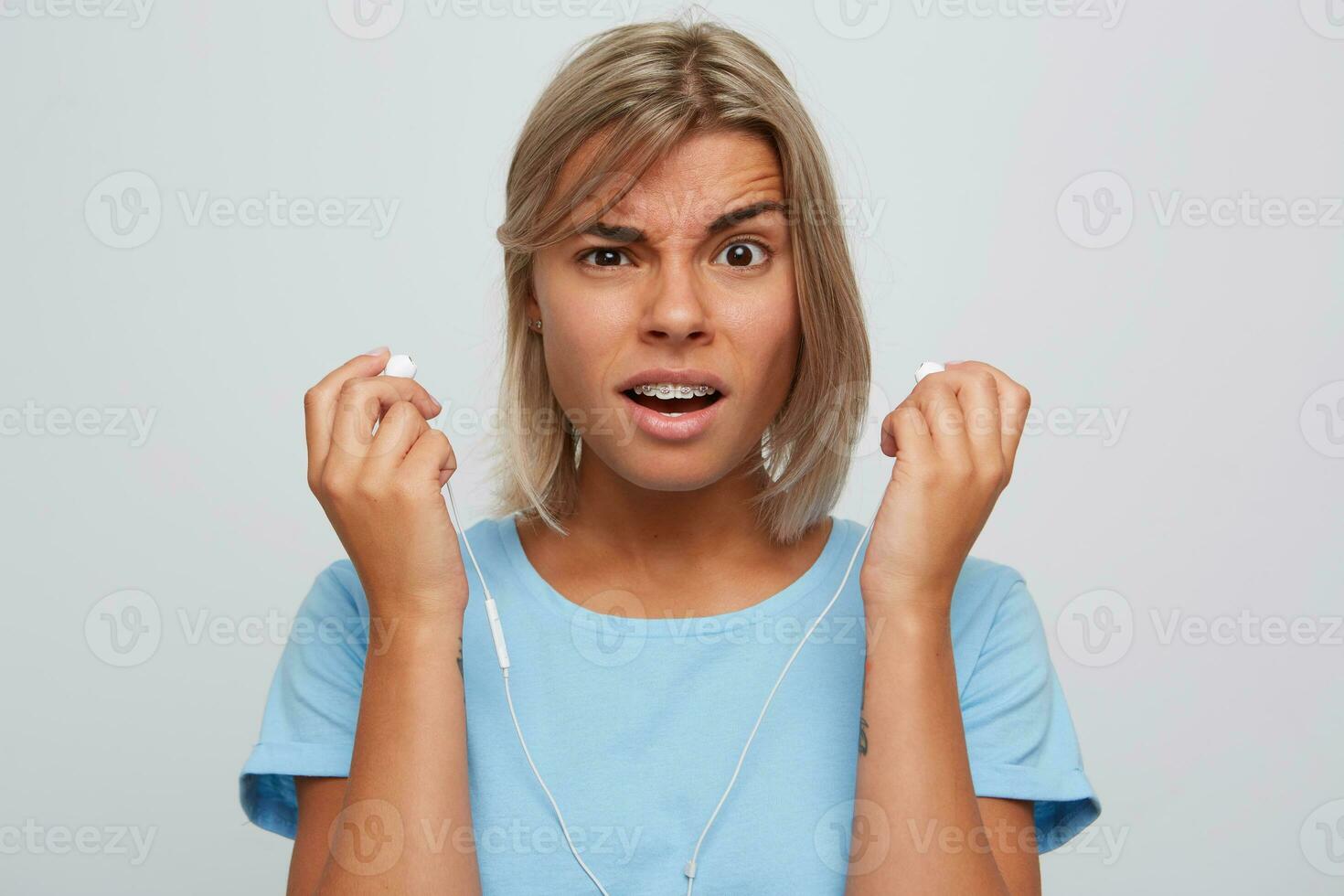 Portrait of confused beautiful blonde young woman with braces on teeth wears blue t shirt looks embarrassed and holding earphones isolated over white background photo