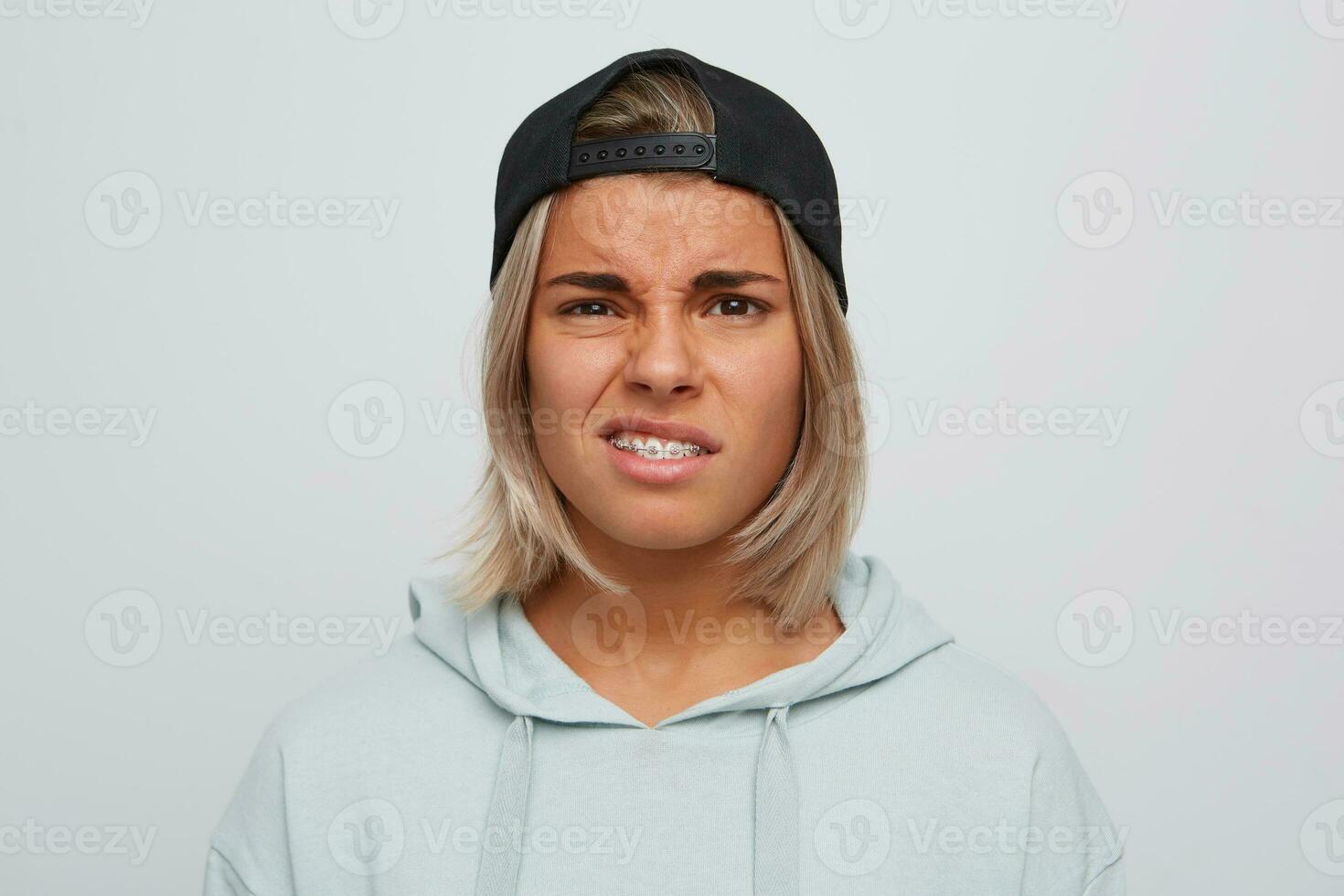 Closeup of sad unhappy blonde young woman with braces on teeth wears black cap and hoodie looks upset and displeased isolated over white background photo