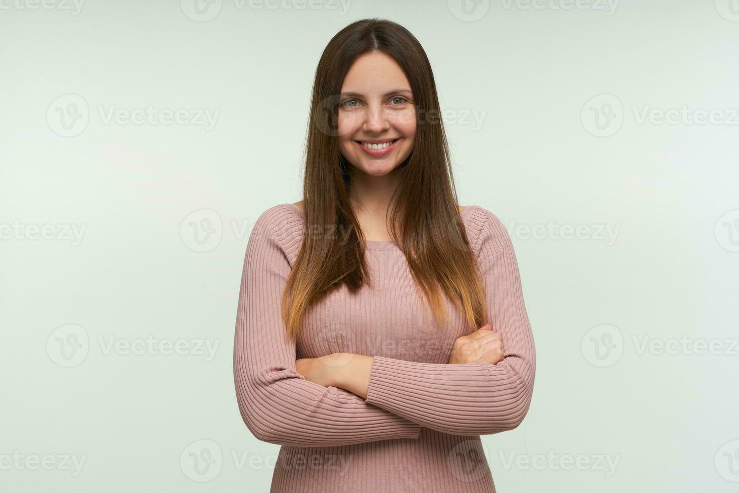 Portrait of young woman with long hair down, stands with hands crossed, looks glad, smiling, dressed in a tight pink knit sweater, isolated over white background photo