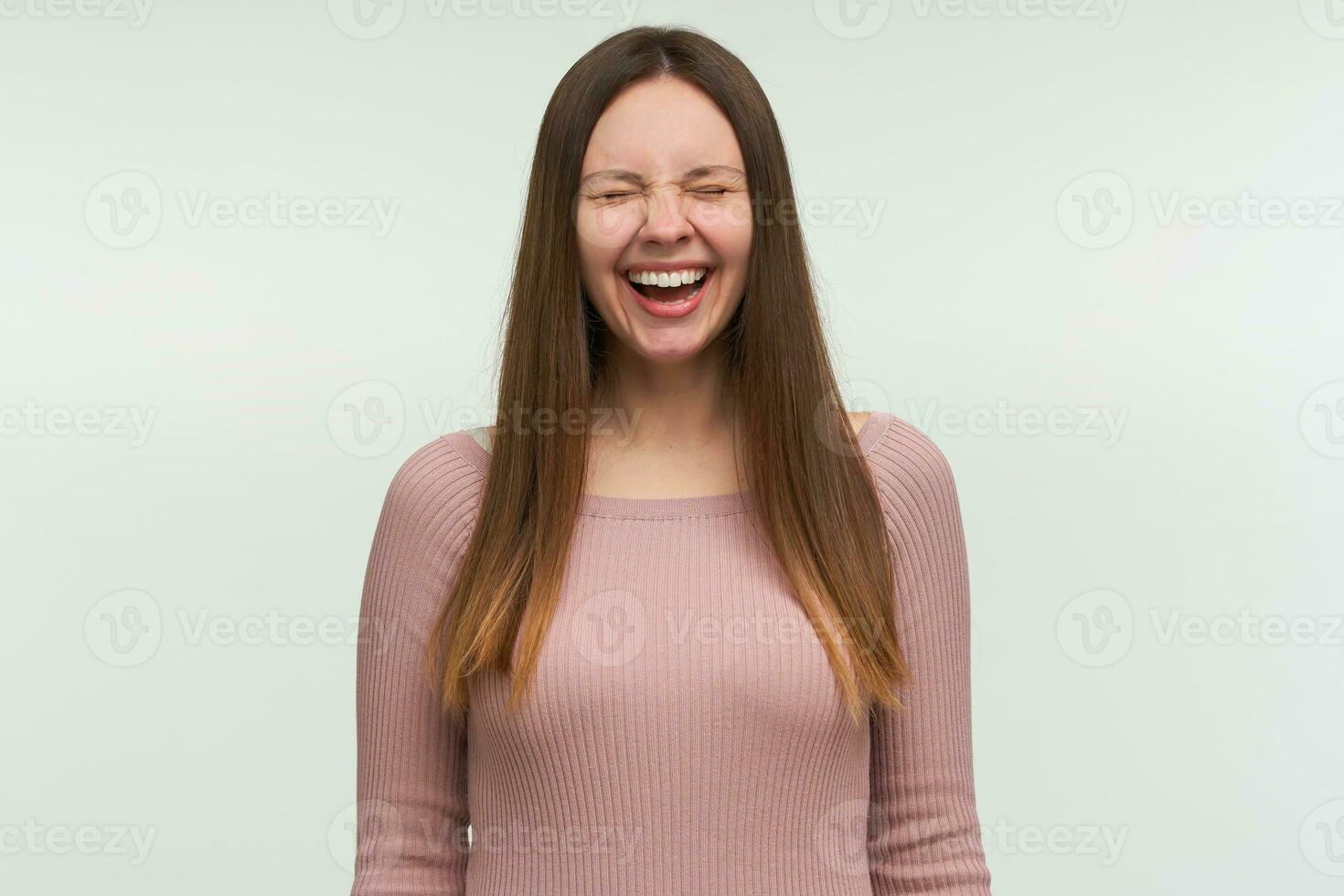 Young woman laughs at a good joke, looks funny, amusing, cheerful, closes eyes with happiness and toothy smile, dressed in a tight pink knit sweater, isolated over white background photo