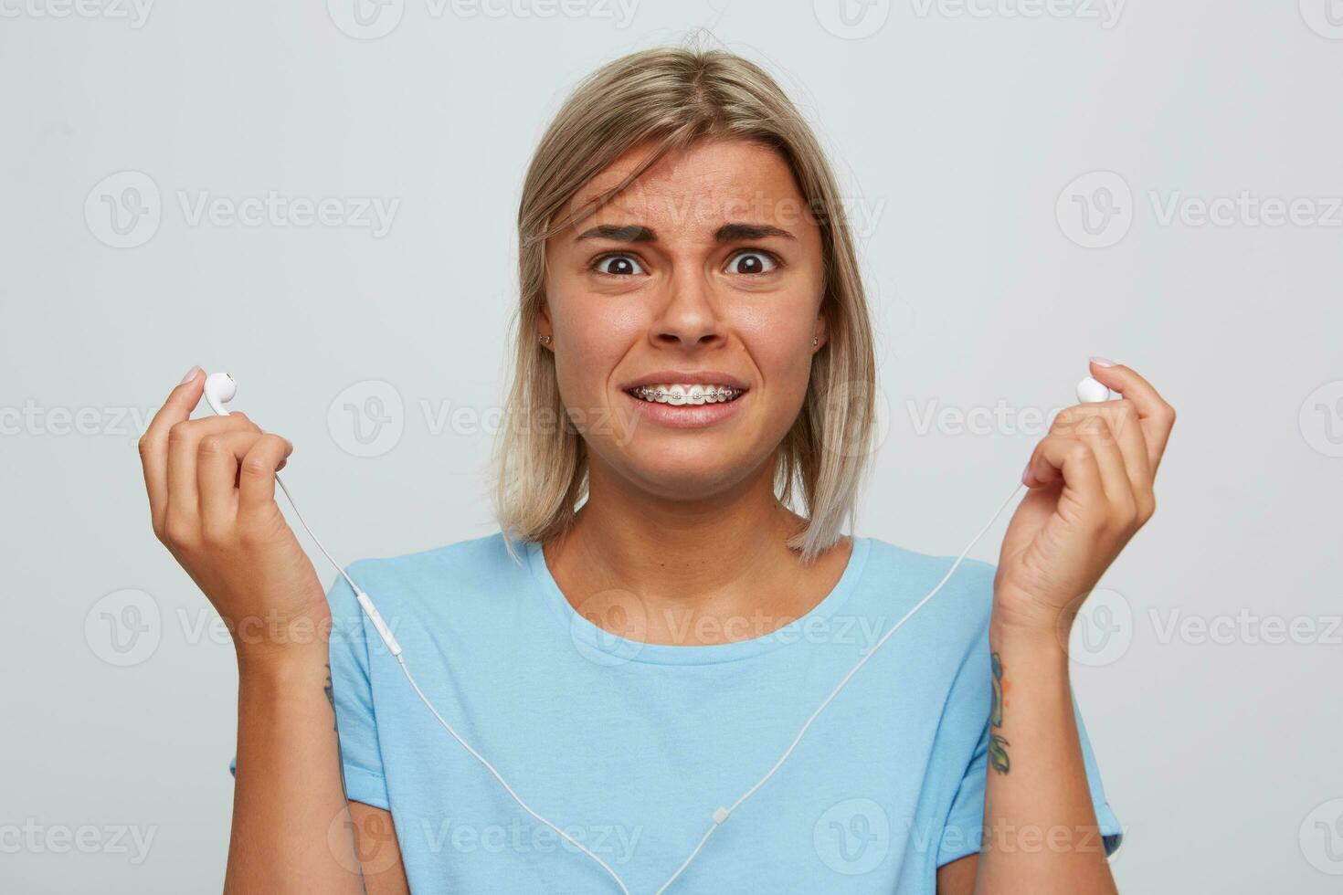 Closeup of shocked confused blonde young woman wears blue t shirt holding earphones, looks embarassed and listening to music isolated over white background photo