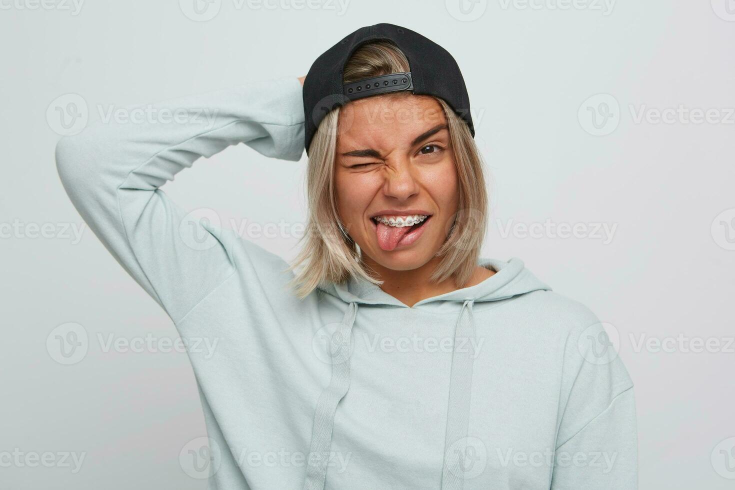 Portrait of funny relaxed blonde young woman with braces on teeth wears black cap and hoodie shows tongue, winking and having fun isolated over white background photo