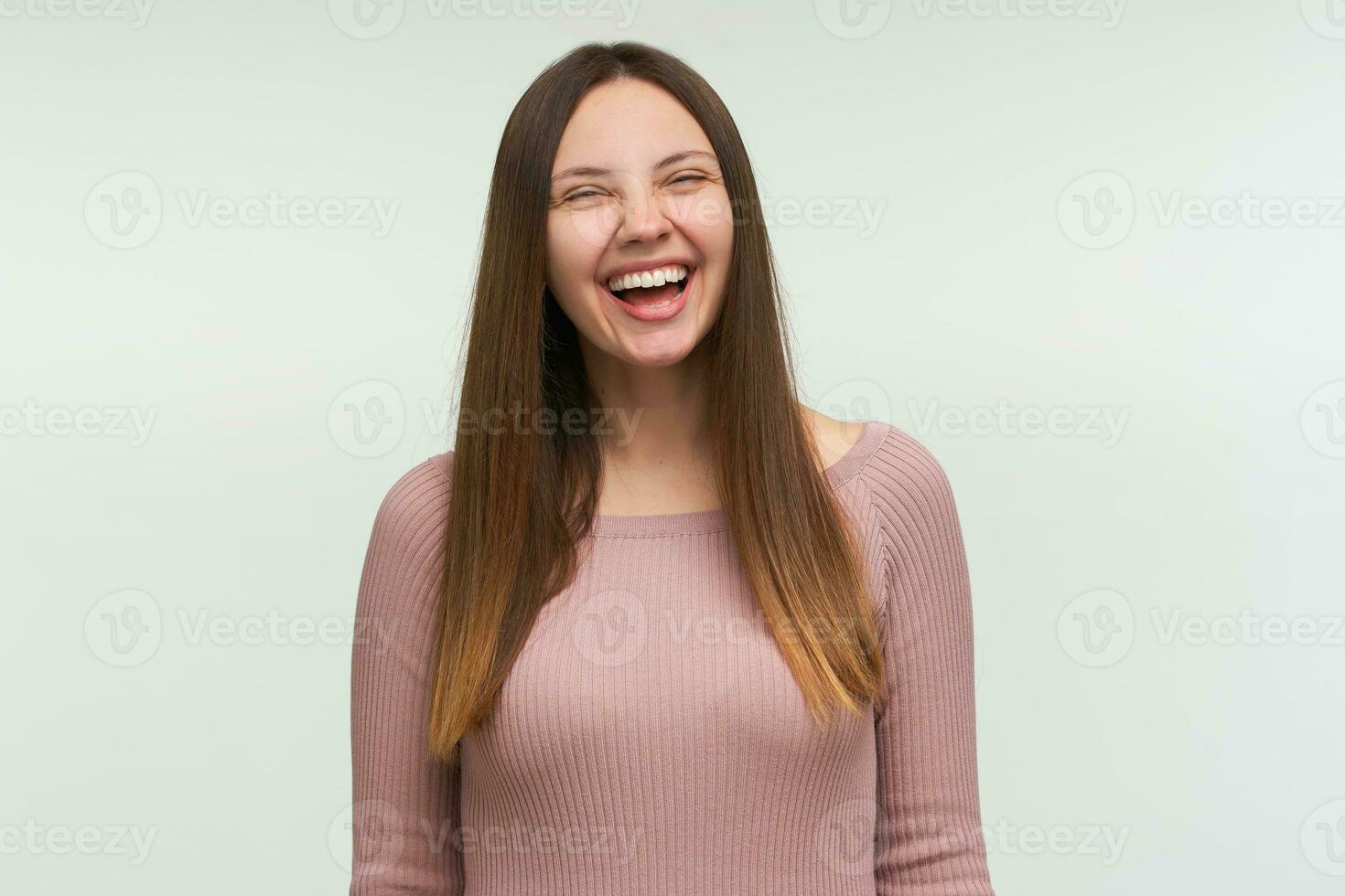 riendo joven mujer con largo pelo abajo, mira divertido, divertido, alegre, se ríe atornillar arriba su ojos, vestido en un apretado rosado tejer suéter, aislado terminado blanco antecedentes foto