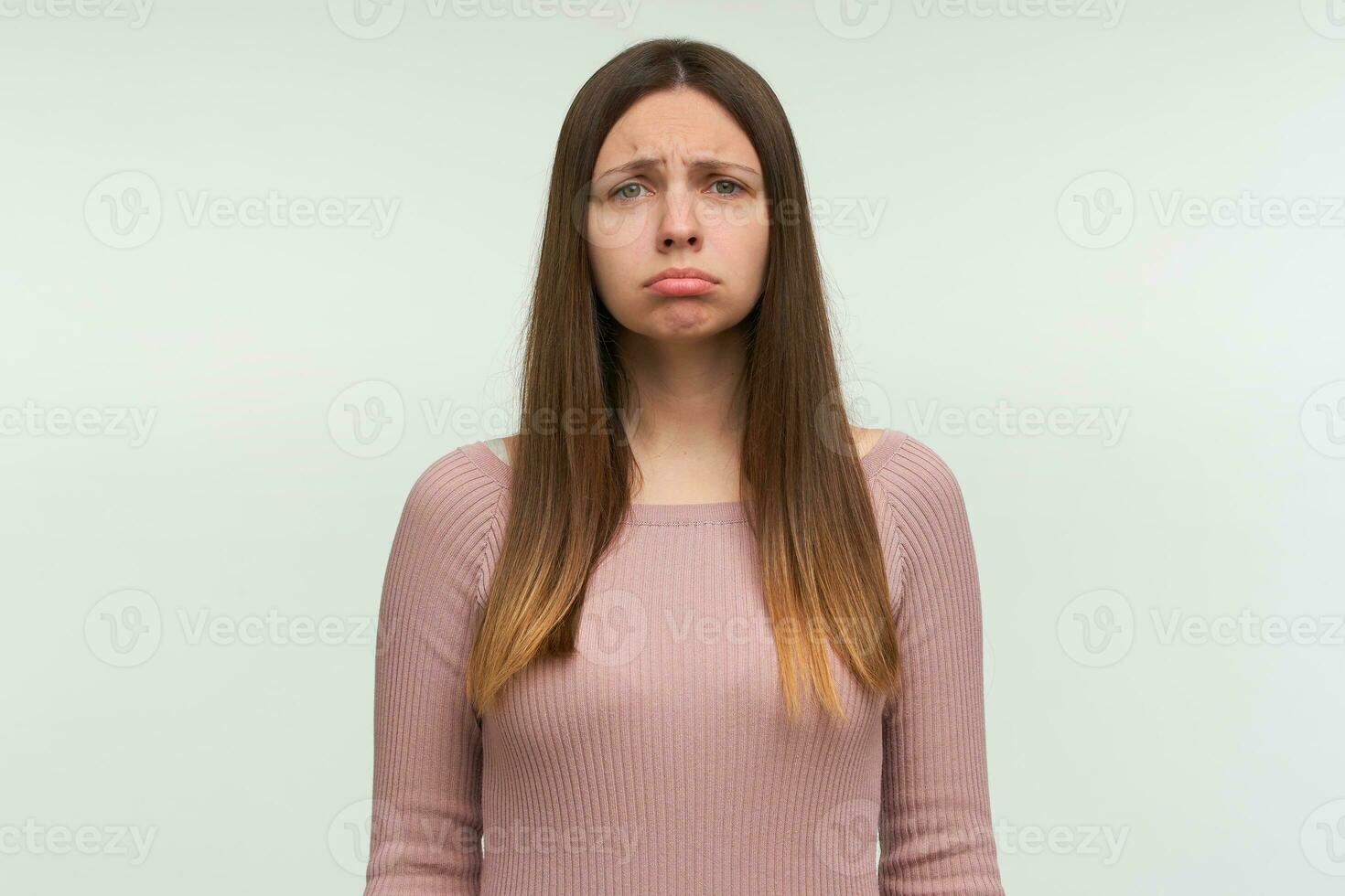 Dissatisfied young female purses lower lip, being abused by something unpleasant, has unhappy expression, dressed in casual pink knit sweater, stands indoor against white wall photo