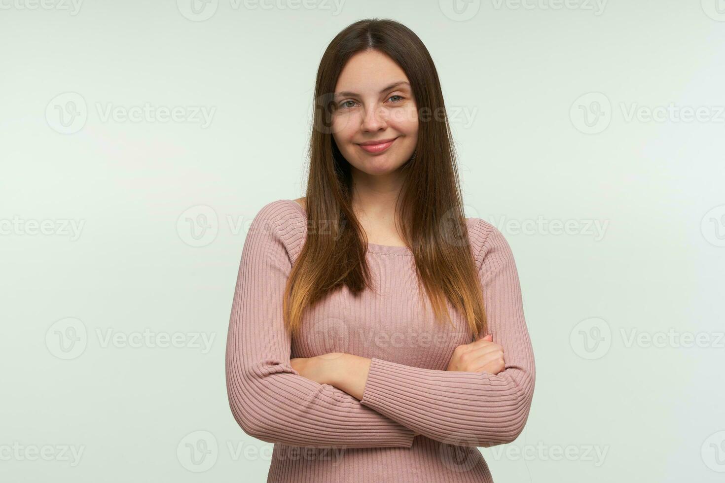 Young woman stands thoughtful, with a sly smile, considering a cunning plan, with hands crossed, looks joyful, dressed in a tight pink knit sweater, isolated over white background photo