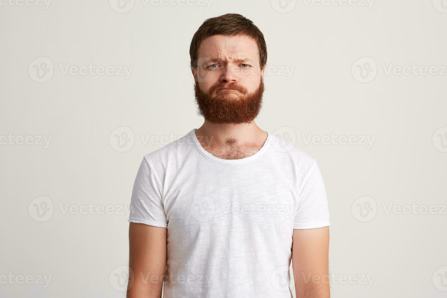 Portrait of serious strict bearded young man wears t shirt standing and feels worried isolated over white background Looks directly in camera photo