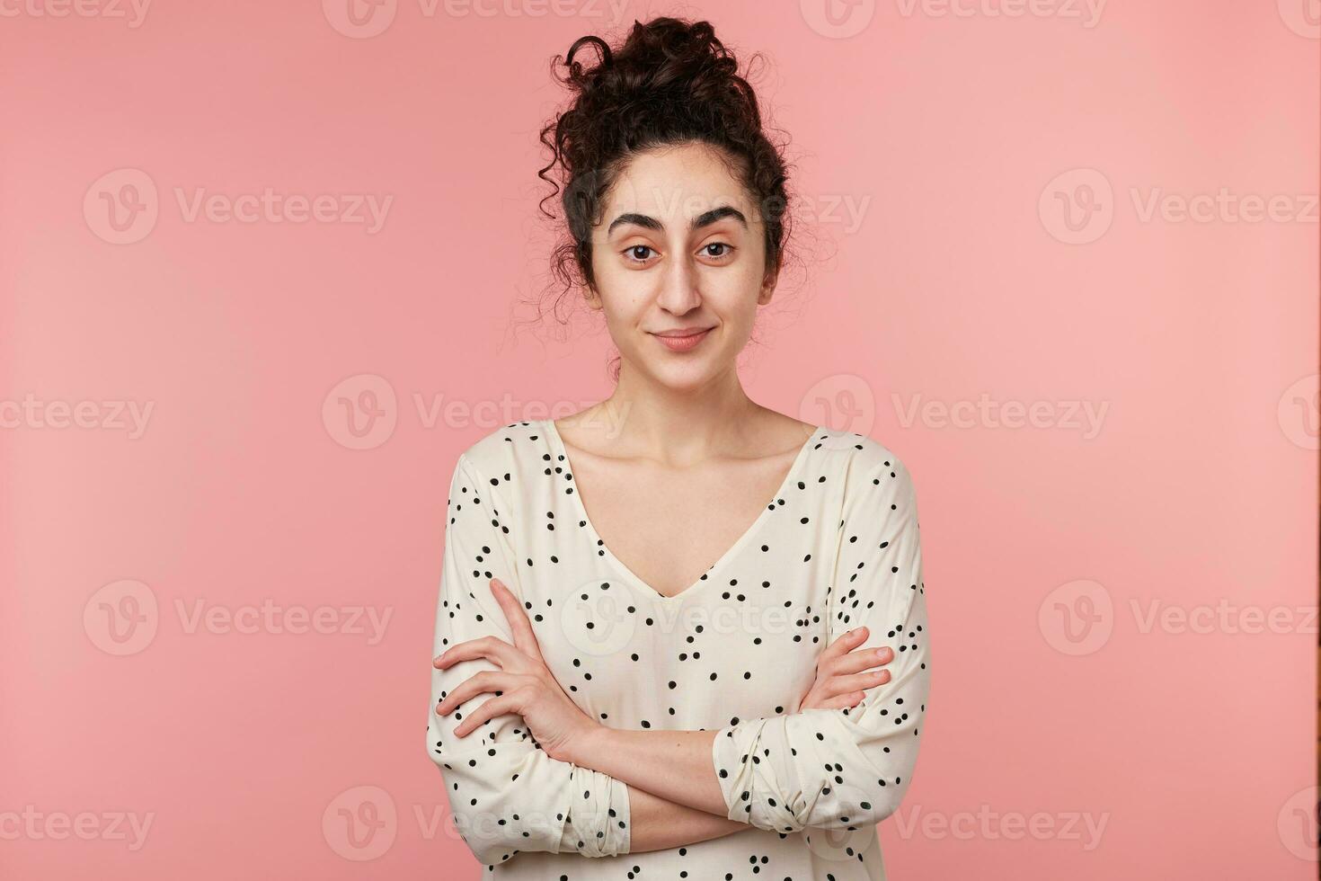 seguro de sí mismo joven pelinegro Rizado niña en pie con cruzado brazos, mirando con interesar y curiosidad, vestido en blusa con polca puntos, aislado en rosado pared foto