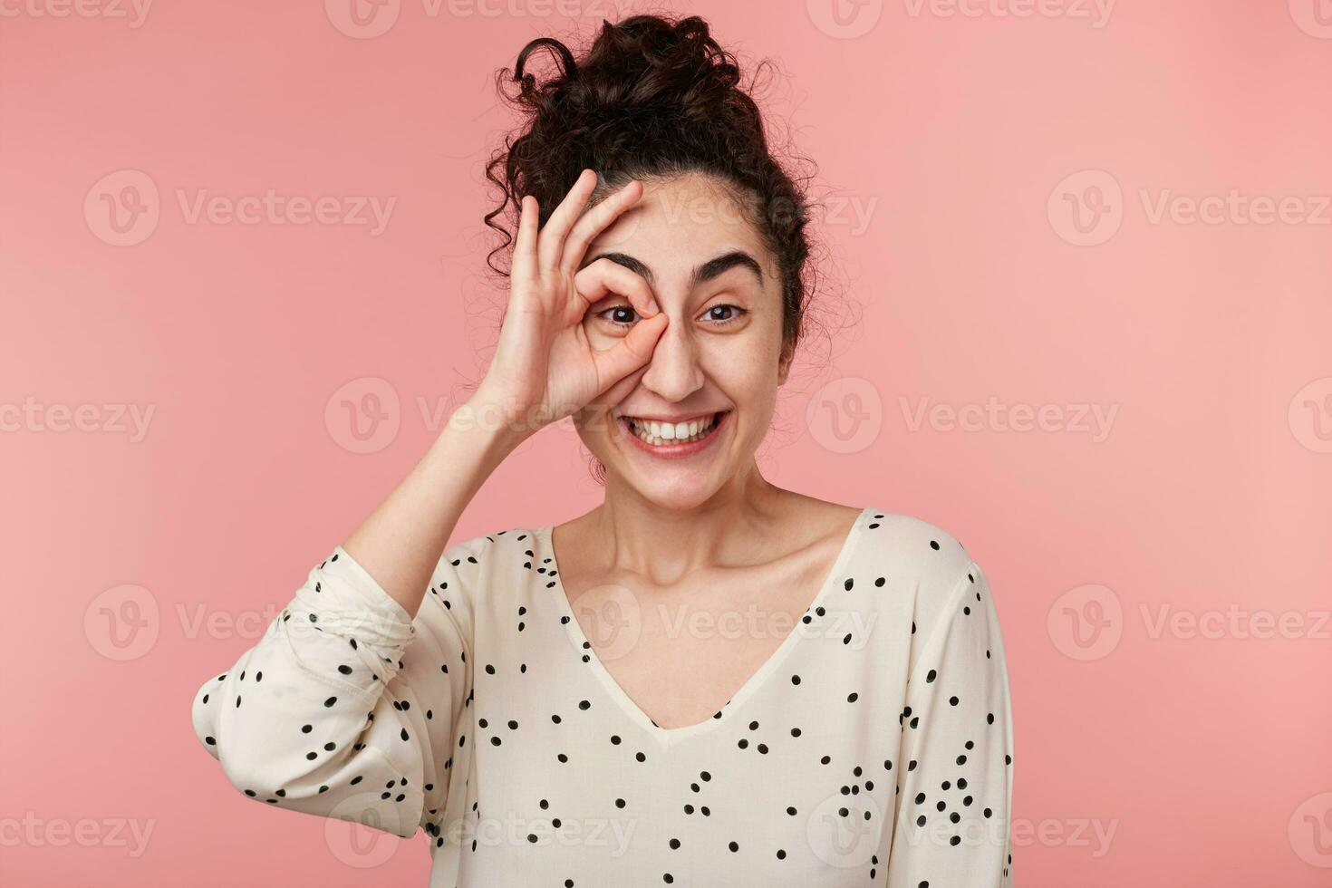 Indoor portrait of funny cute attractive brunette girl with collected wave hair making okey symbol close to eye with one hand, widely smiling, in blouse with polka dots,isolated on pink background photo