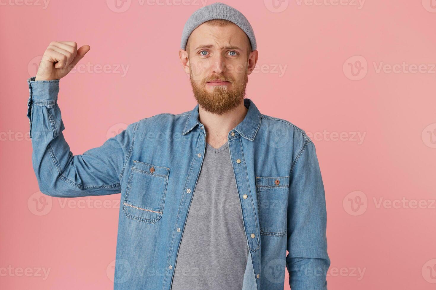 A young bearded guy can not boast of his strength, displeased with his physical form, raised his bent arm to demonstrate weak muscles, with chagrin looking at the camera, dressed in a denim shirt photo