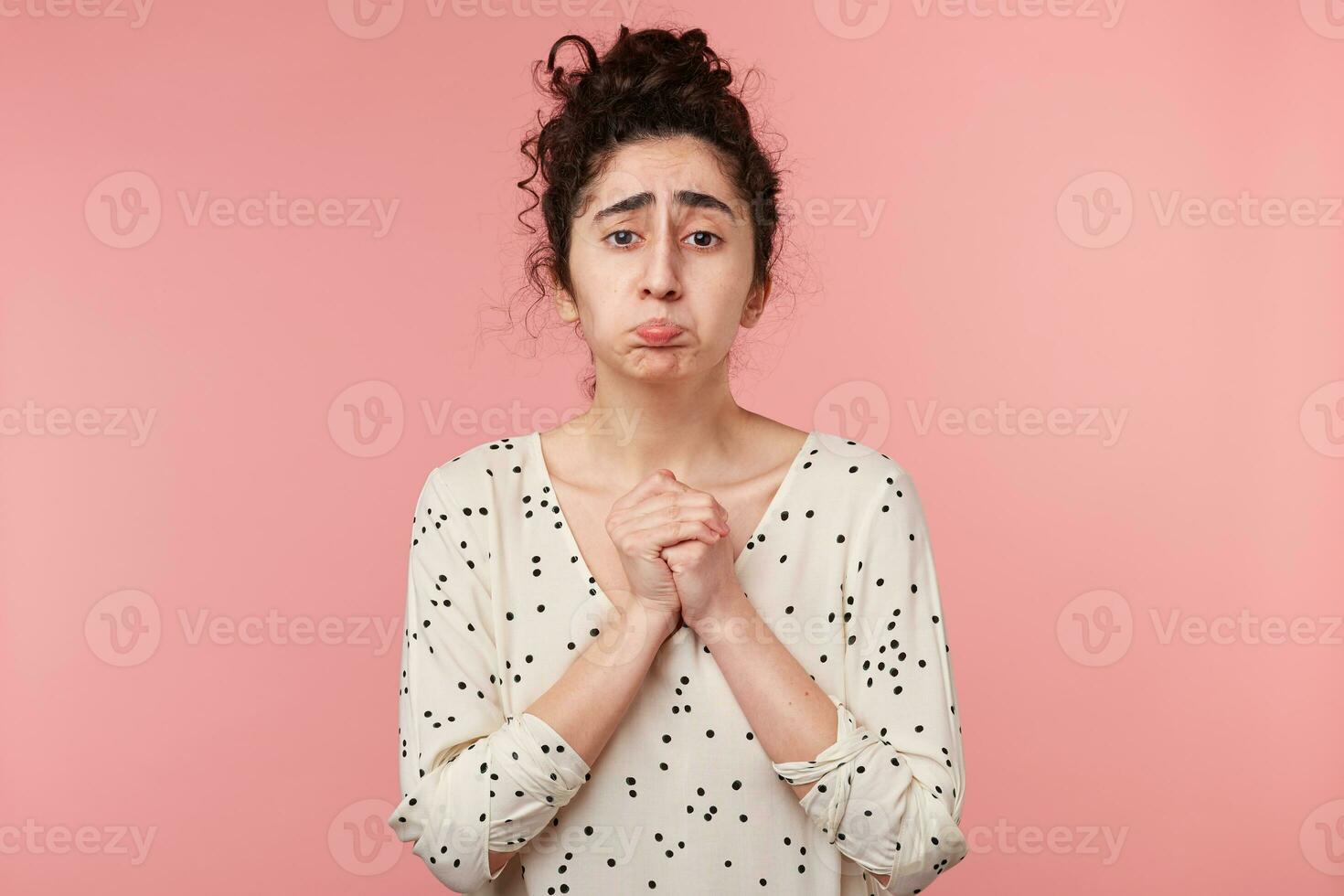 Brunette girl imploring begging, pouted her cheeks, looks sad cherish hope, folded hands in a praying gesture, dressed in blouse with polka dots, going to cry, isolated on pink photo