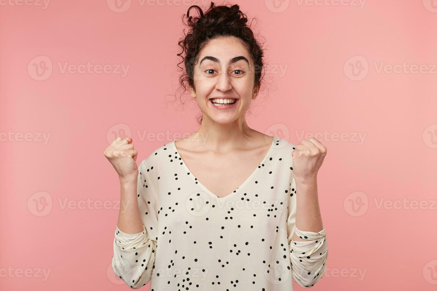 alegre joven Rizado morena niña con marrón ojos en blanco blusa con polca puntos elevado manos y apretado puños en emoción cuando tiene el victoria y se convirtió un ganador en competencia, terminado rosado pared foto