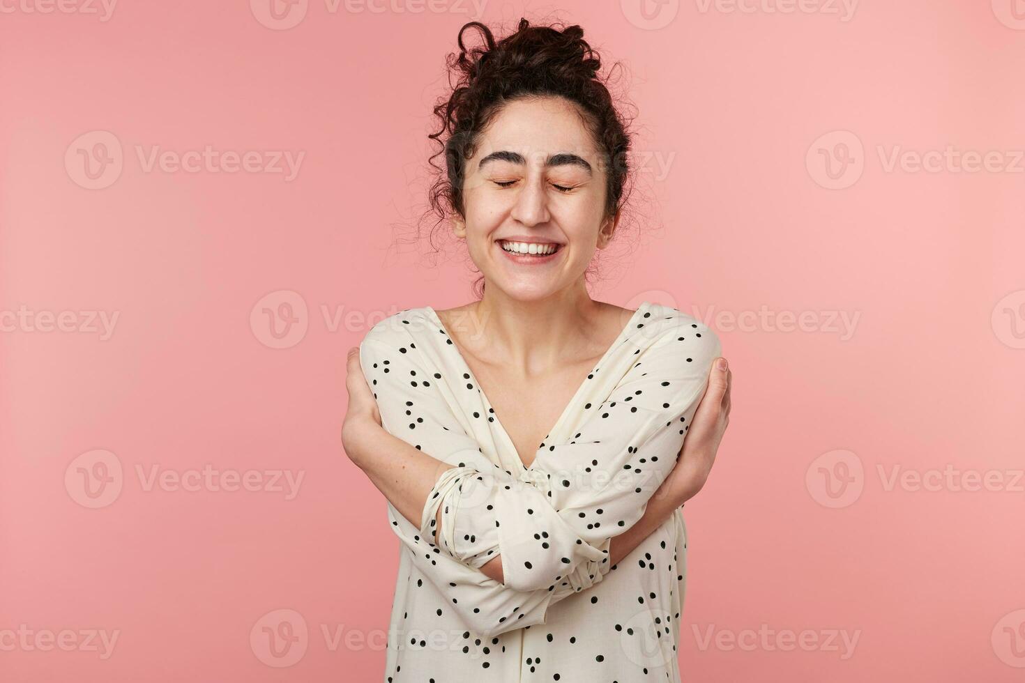 Happy young curly brunette girl in white blouse with polka dots closed eyes and hugs herself in excitement and happiness when got the victory or won the main prize in draw, over pink background photo