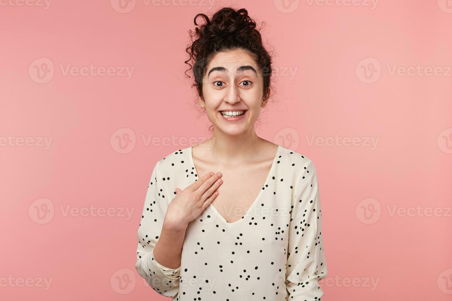 Young cheerful attractive curly brunette girl with brown eyes in white blouse with polka dots pointing with hand at herself in excitement being chosen for the main role in university play photo