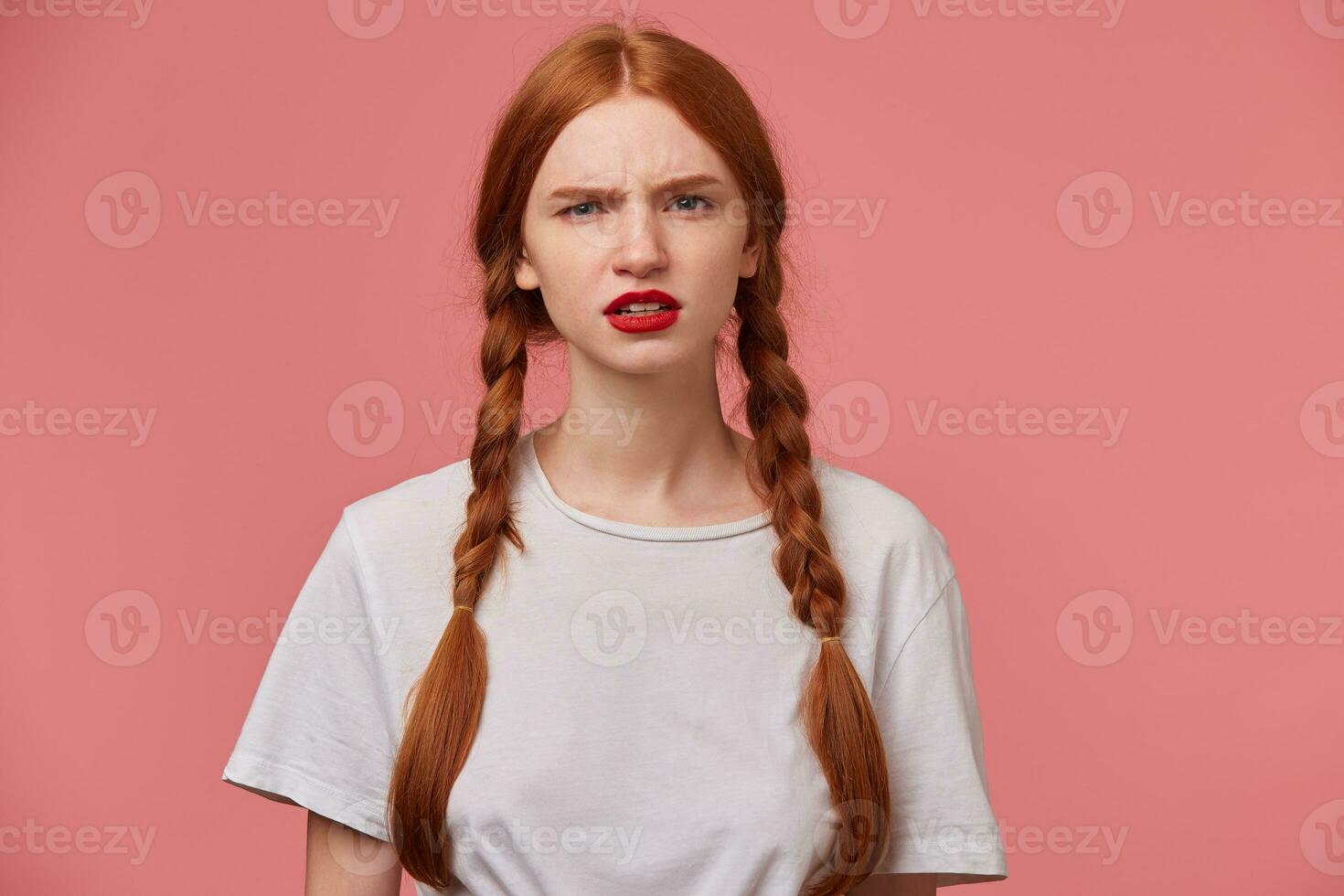 Indoor portrait of young ginger female with freckles posing over pink background looking into camera with sad facial expression photo