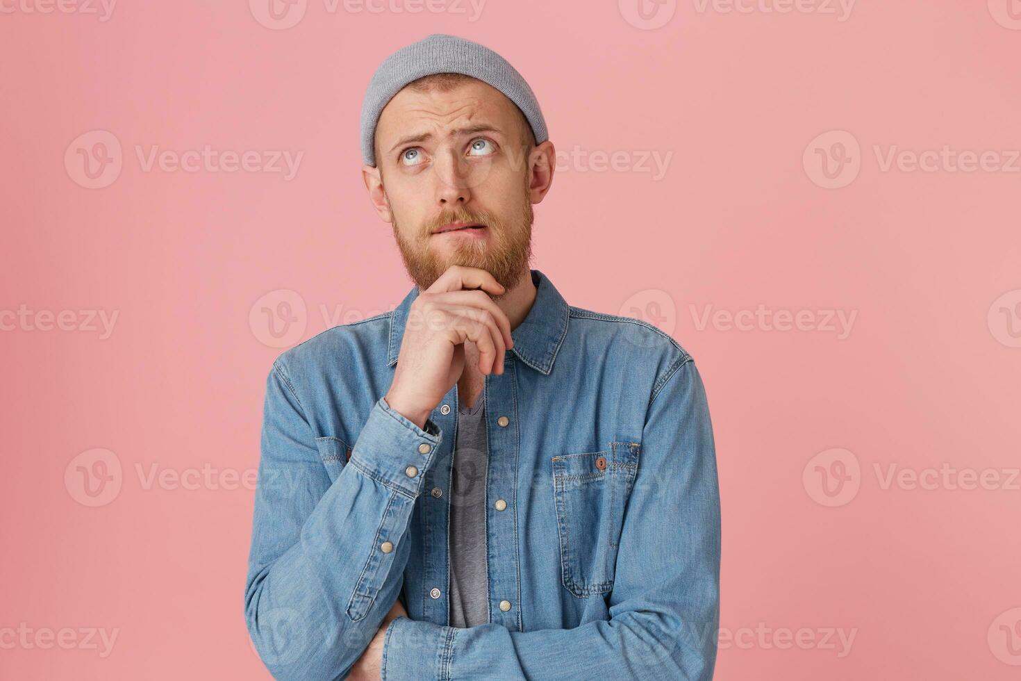 Bearded man is focused, keeps fist near the chin, trying to make a decision, looking upwards with thoughtful sad look, bites his lip, doubts,ponder about something, isolated over pink background photo