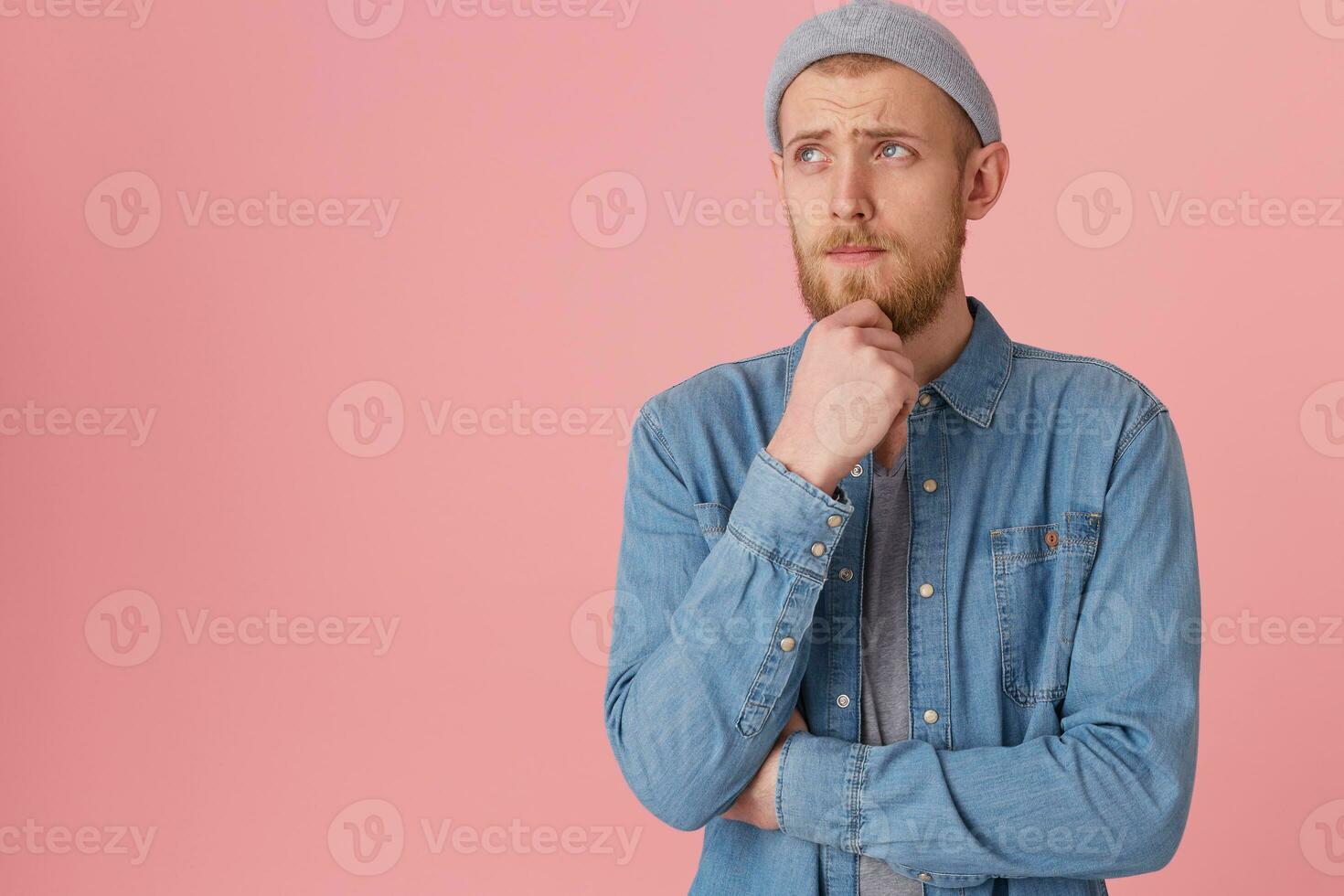 Young man is focused, keeps fist near the chin trying to remember something, looking away with sad thougtful look, ponder about something, isolated over pink background with copy space photo