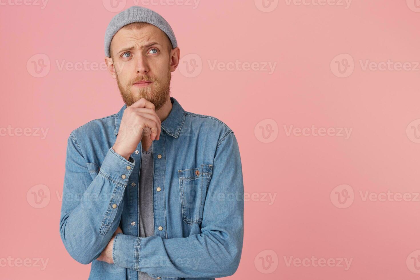 Young man thinks, holds his beard, looking away with sad thougtful look, ponder about something, isolated over pink background with copy space for your text photo