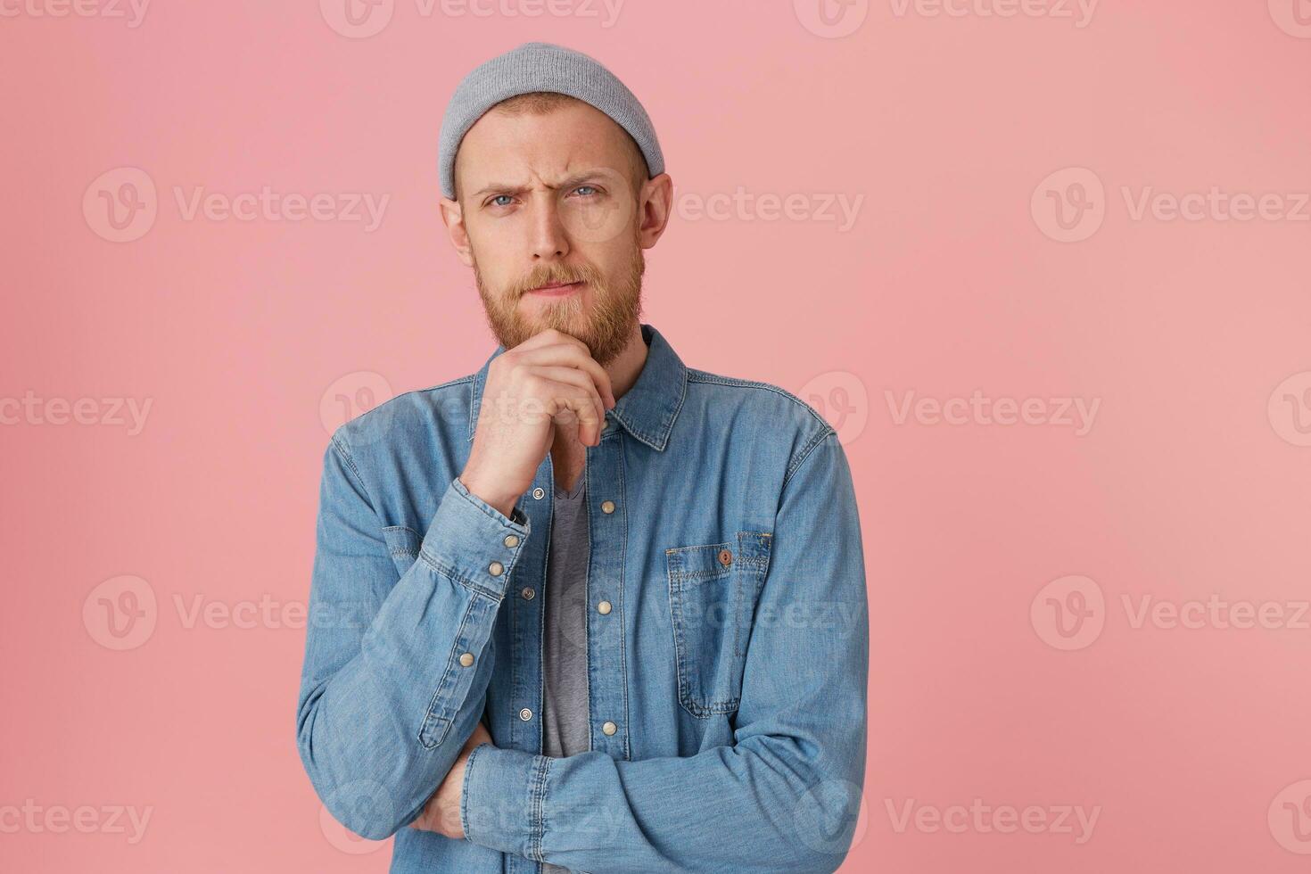 Serious attentive bearded man keeps fist near the chin, looking camera with thoughtful suspicious mistrustful look, doubts, ponder about something, isolated over pink background photo