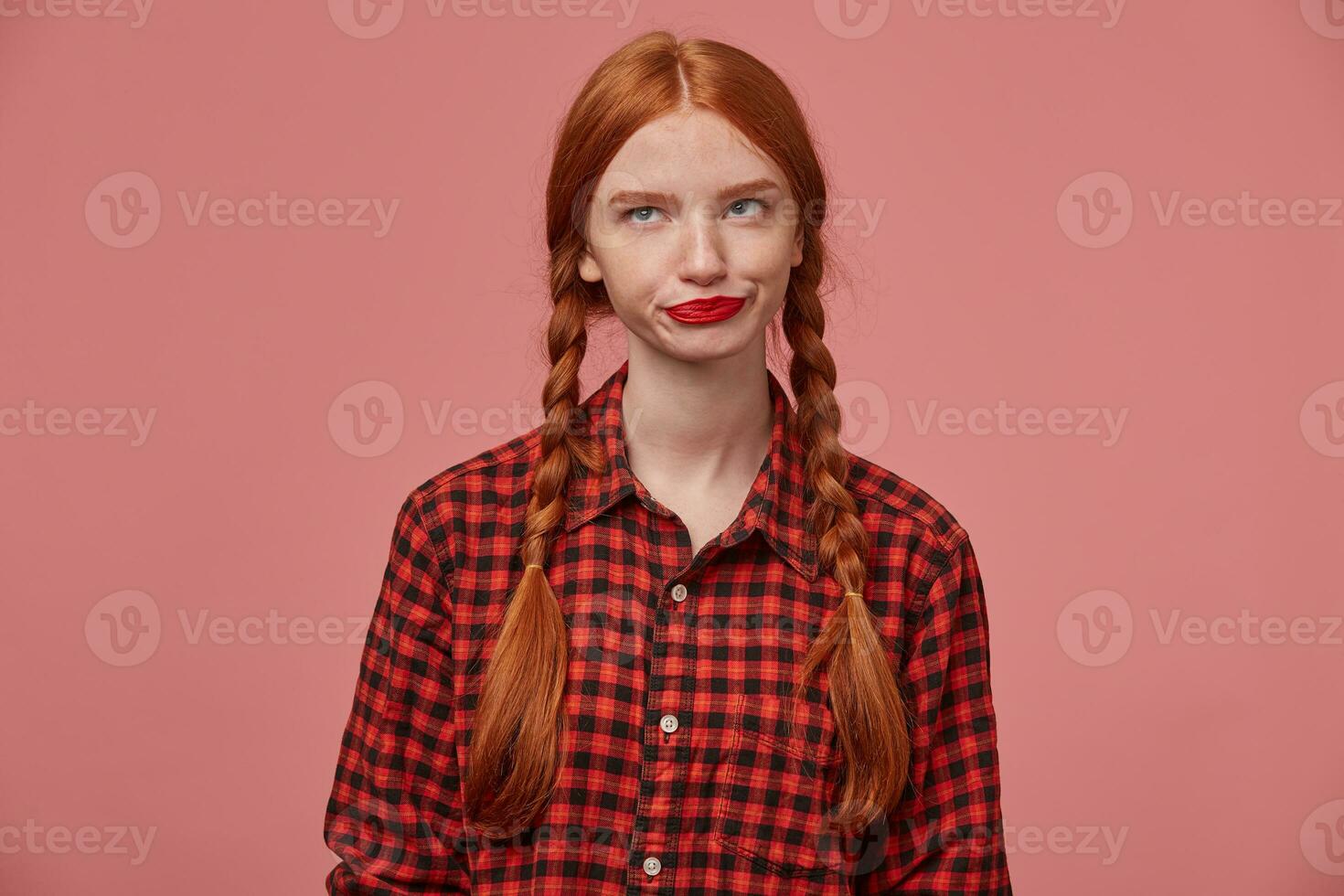 indoor shot of sad negative ginger female, pressing her lips, looks aside at copy space with confused facial expression photo
