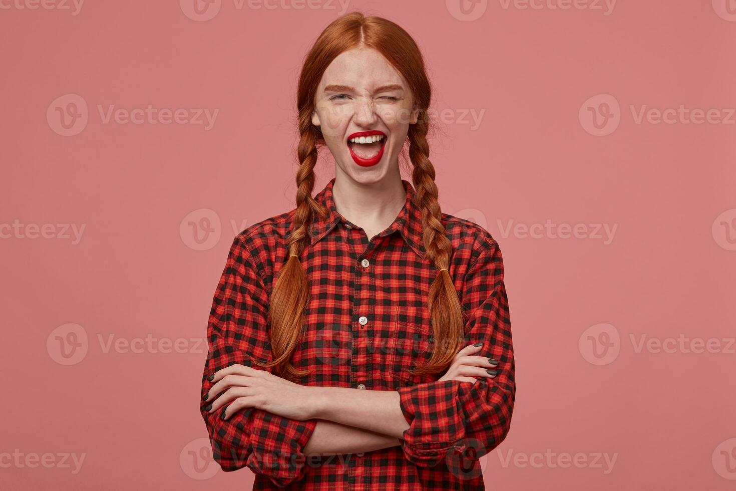indoor shot of young redhead teenage girl, keeps her hands crossed, smiles broadly and wink with positive facial expression. Isolated over pink background photo