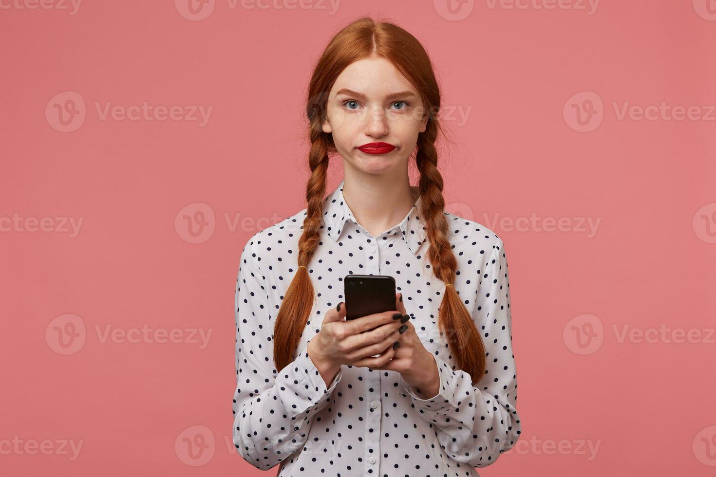Indoor portrait of young ginger female with freckles posing over pink background holding phone with sad depressed facial expression photo