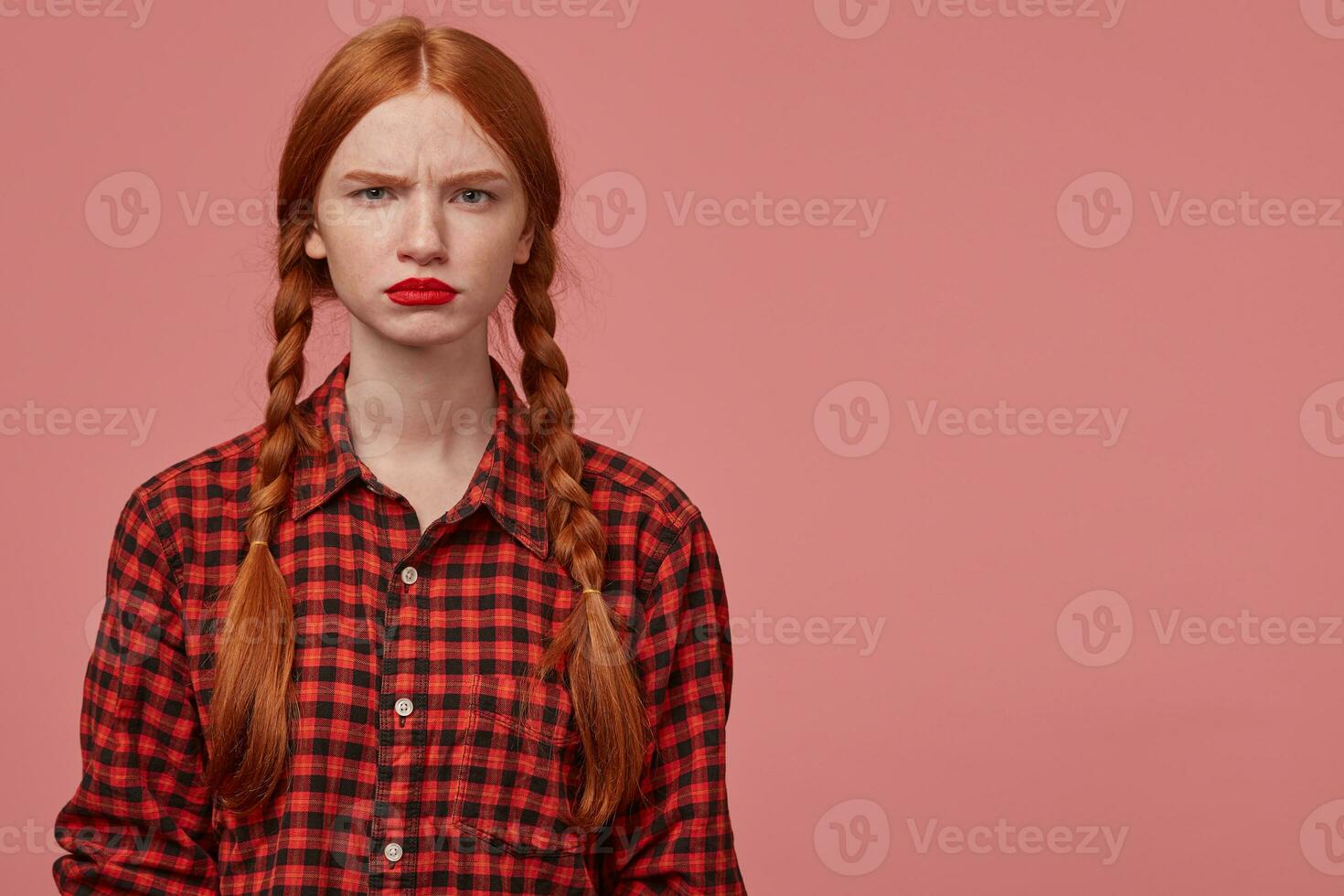 indoor shot of serious and sad young ginger female, pressing her lips and knit eyebrows, starring into camera with confused facial expression. Isolated over pink background with copy space photo