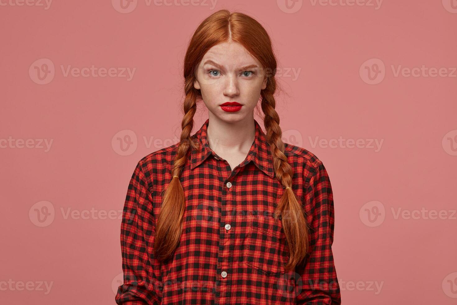 Indoor shot of young ginger female, pressing her lips with negative, mad facial expression, keeps her eyebrow raised and starring into camera. Isolated over pink background photo