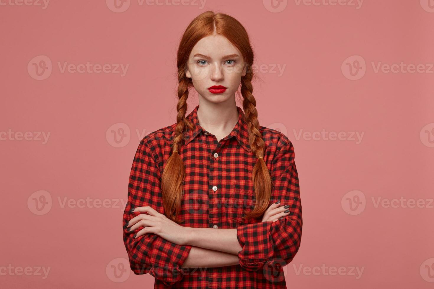 indoor shot of young sad and confused female, keeps her hand crossed on chest, looks directly into camera with negative facial expression, raised her eyebrow. Isolated over pink background photo