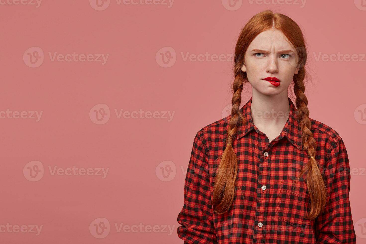 portrait of young ginger female, looks aside at copy space, biting her lips and raised eyebrows with confused facial expression photo