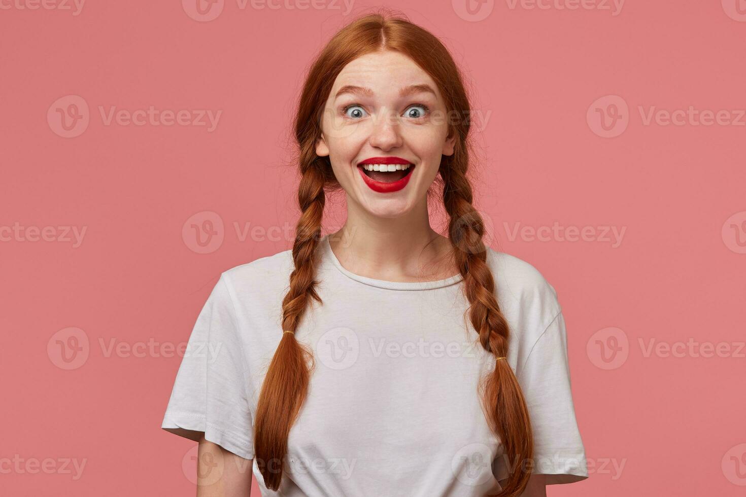 Indoor portrait of young ginger female with freckles posing over pink background looking into camera with shocked, excited facial expression photo