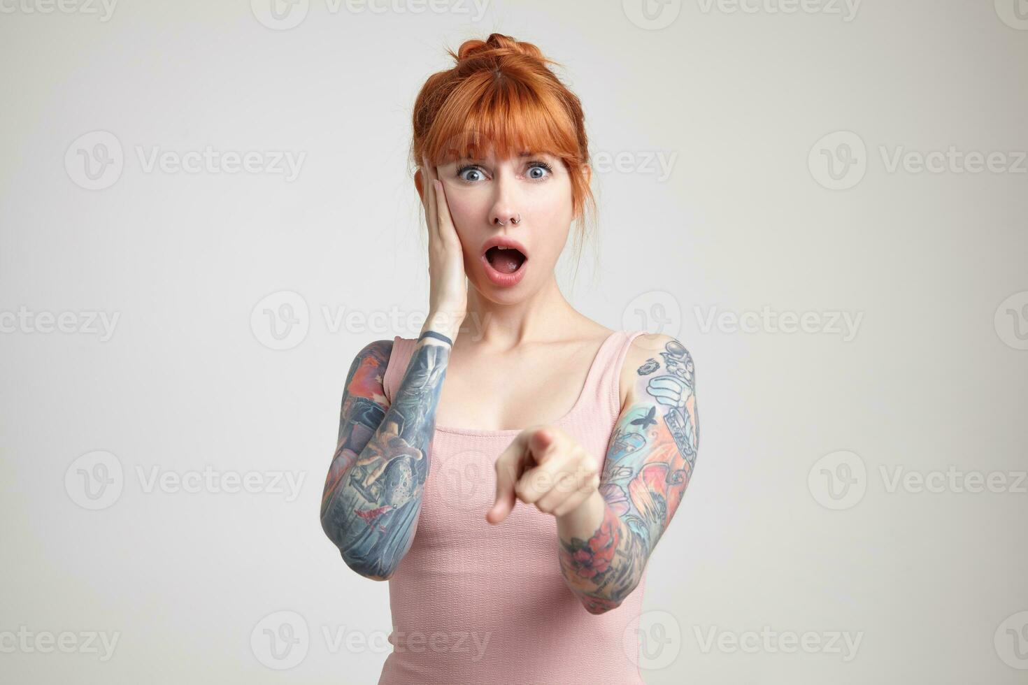 Indoor portrait of young ginger female posing over white wall points into camera with shocked facial expression photo