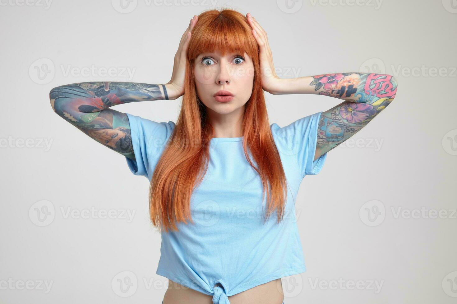 Indoor portrait of young ginger female posing over white wall holding her head and looking into camera with shocked facial expression photo