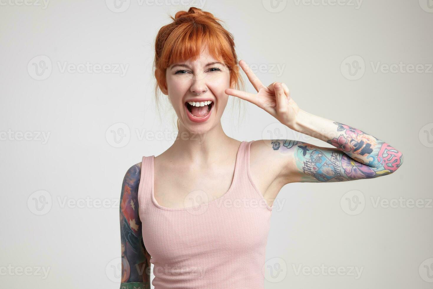 Indoor portrait of young ginger female posing over white wall showing v sign into camera photo