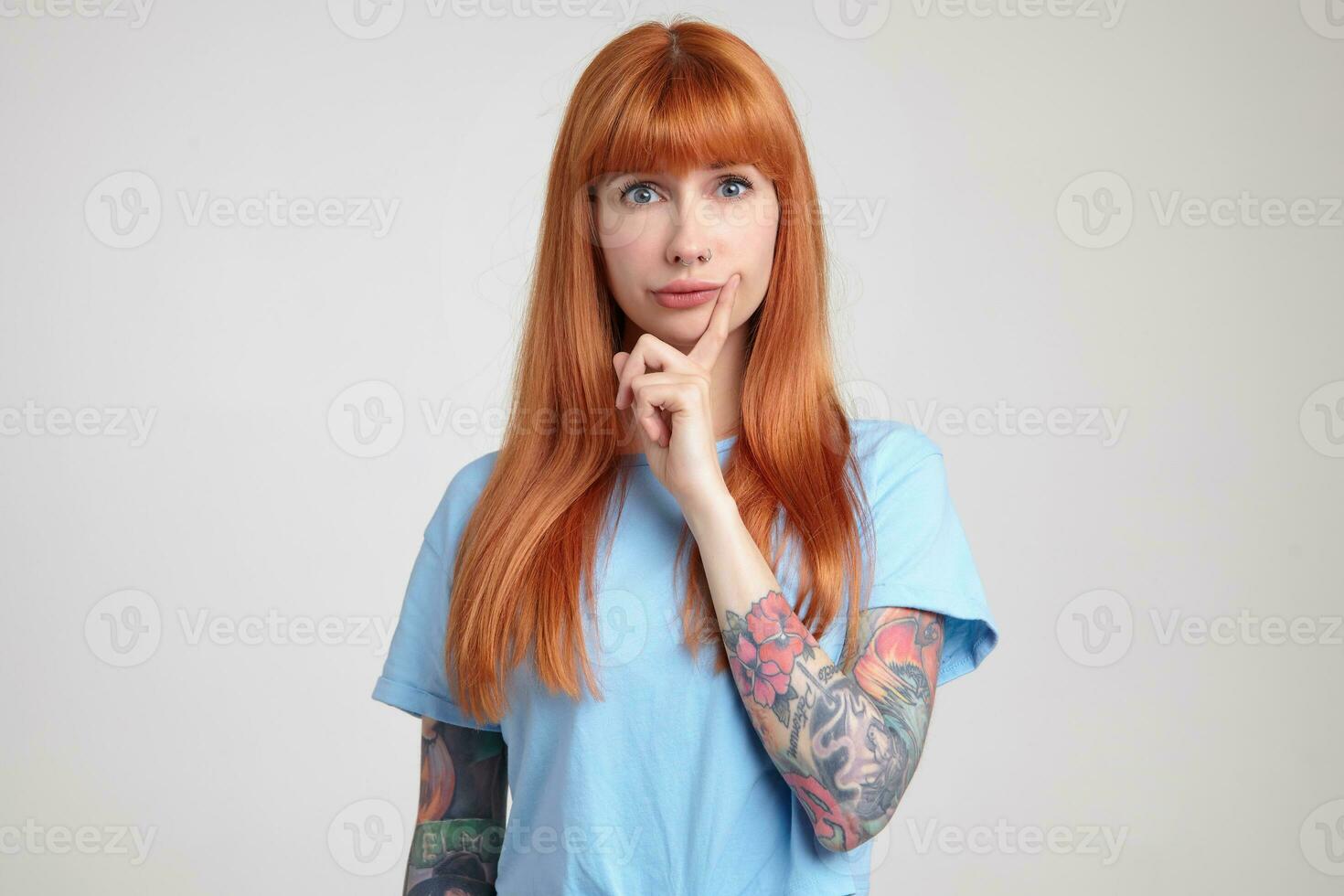 Indoor portrait of young ginger female posing over white wall looking into camera with confused facial expression photo