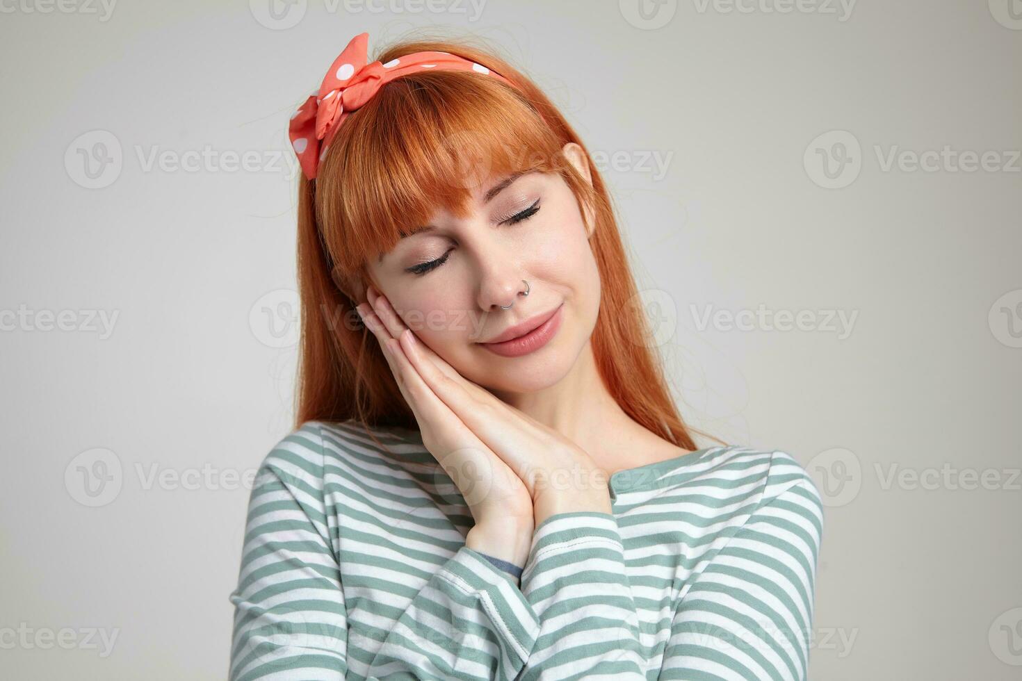 Indoor portrait of young ginger female posing over white wall showing sleep sign into camera photo