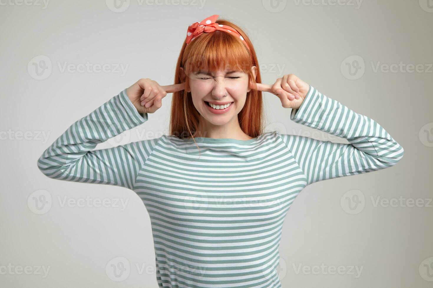 Indoor portrait of young ginger female posing over white wall plug her ears with fingers photo