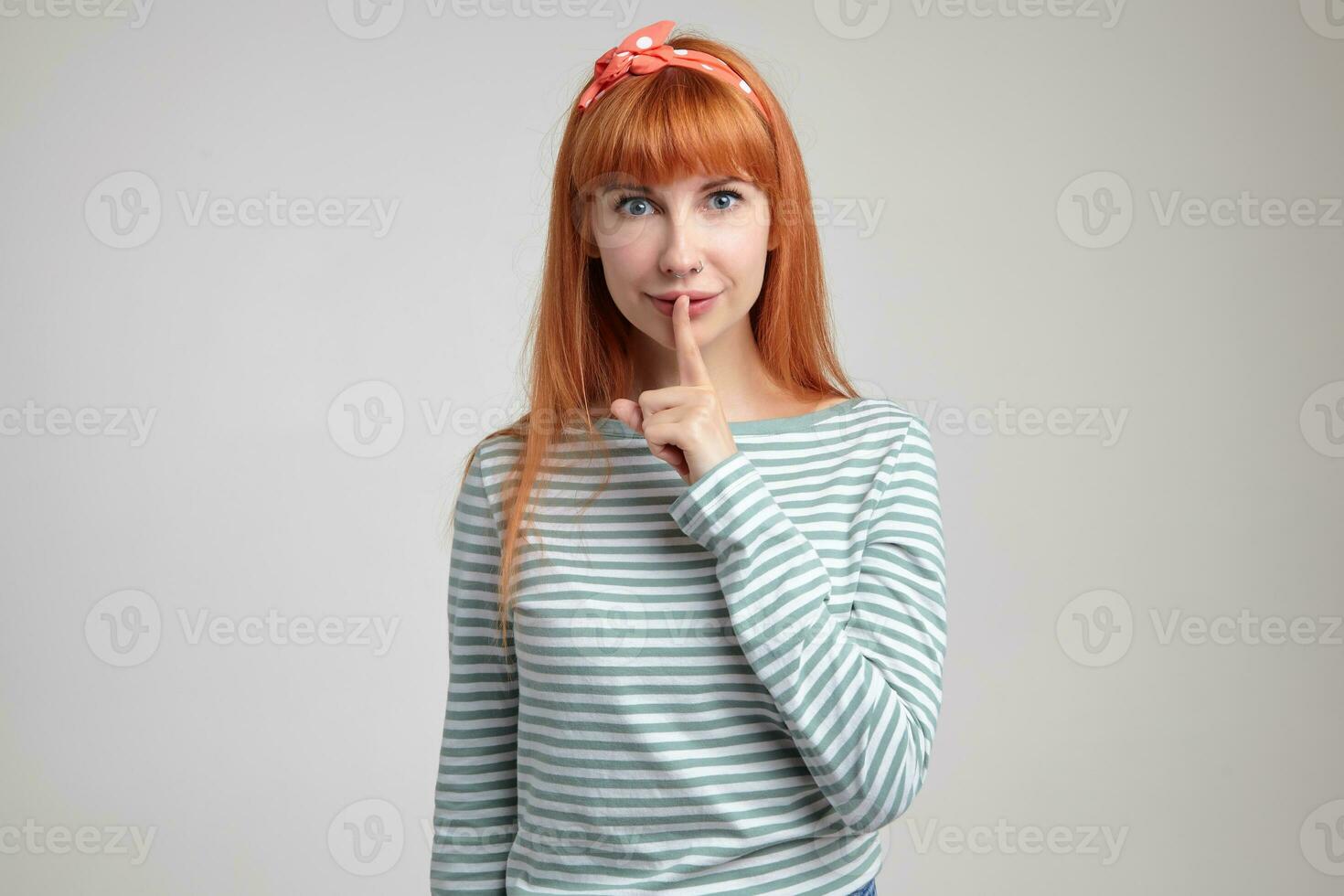 Indoor portrait of young ginger female posing over white wall showing silence gesture into camera photo