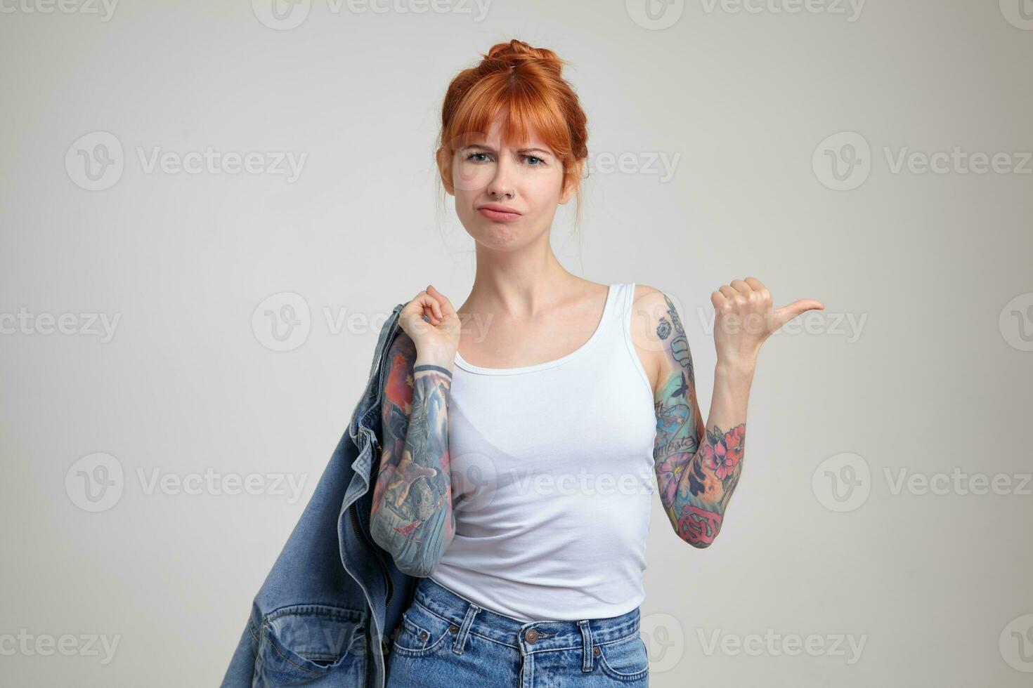 Indoor portrait of young ginger female posing over white wall point with a finger aside at copy space with confused facial expression photo