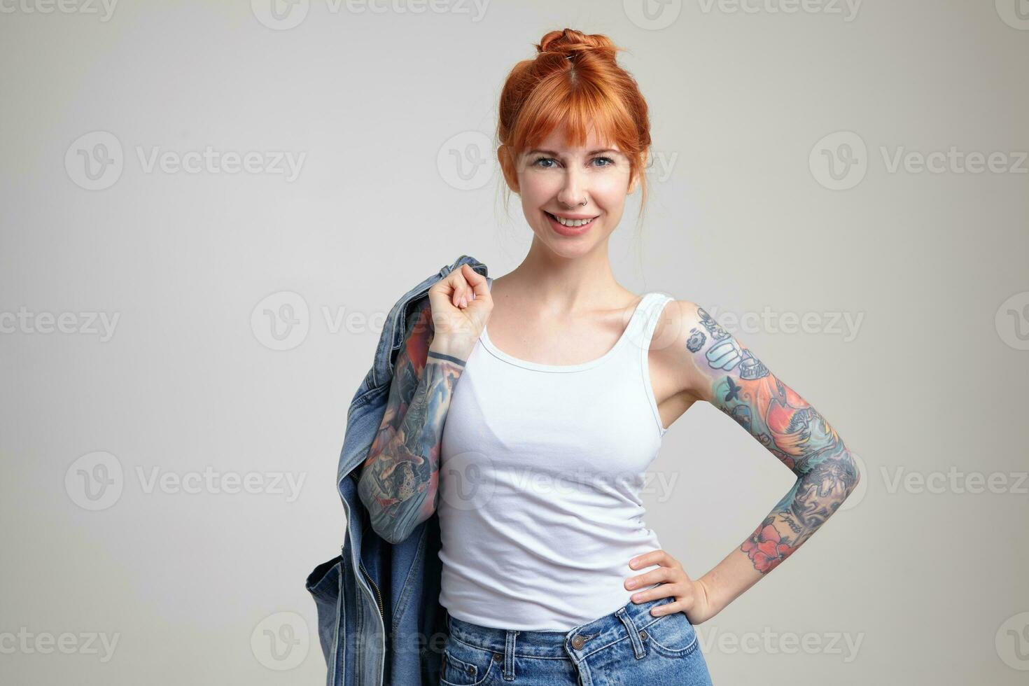 Indoor portrait of young ginger female posing over white wall holding denim jacket and smiles broadly photo