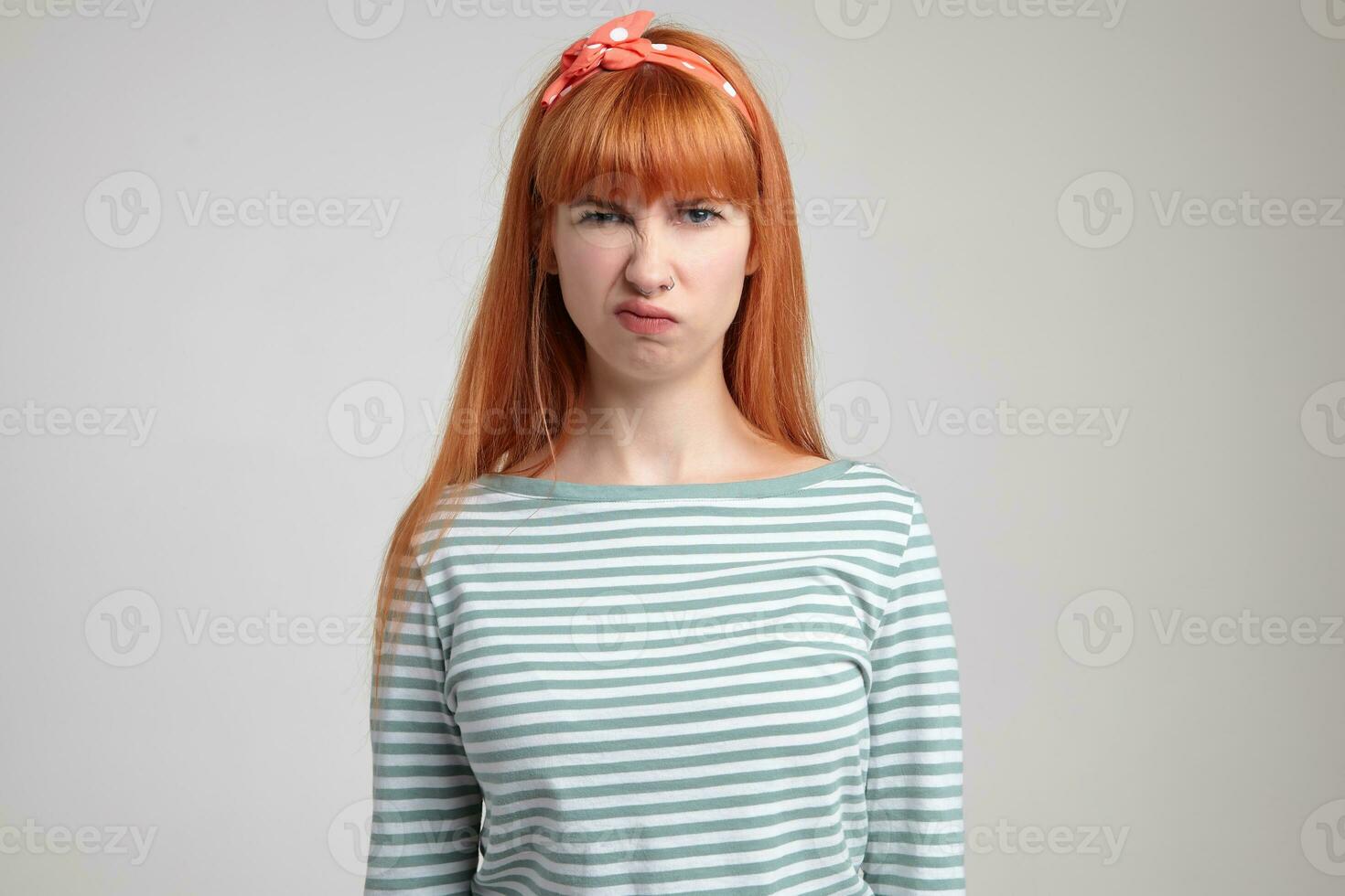 Indoor portrait of young ginger female posing over white wall looking into camera with angry facial expression photo