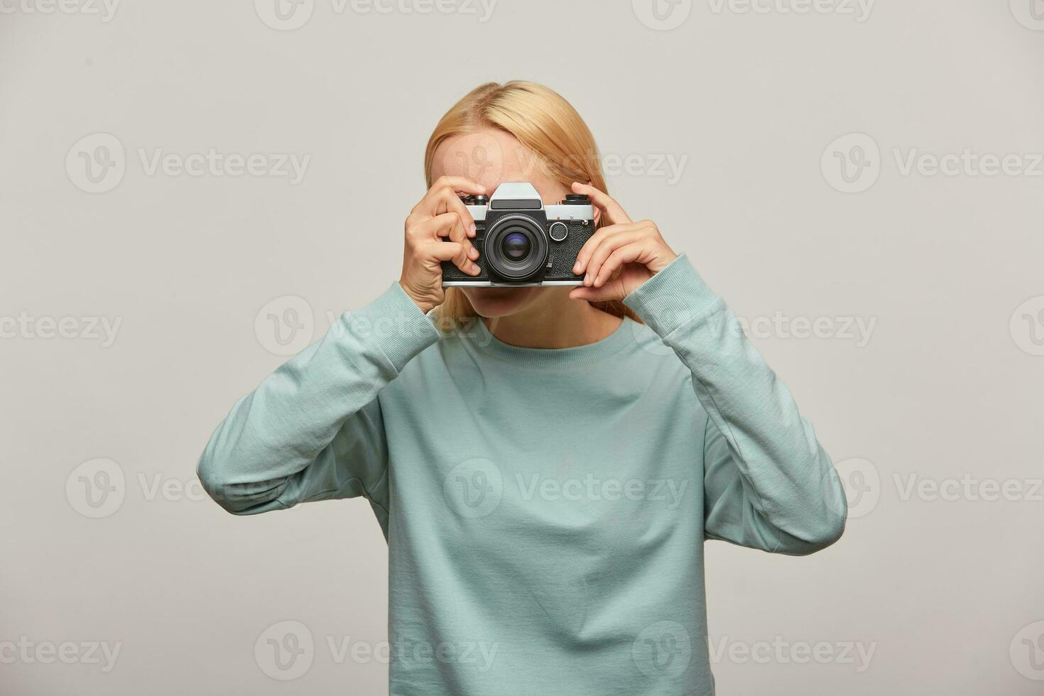 Girl covering her face with the camera making a photo session. Photographer taking photo, working in studio, in-door photography, dressed blue sweatshirt, over grey background