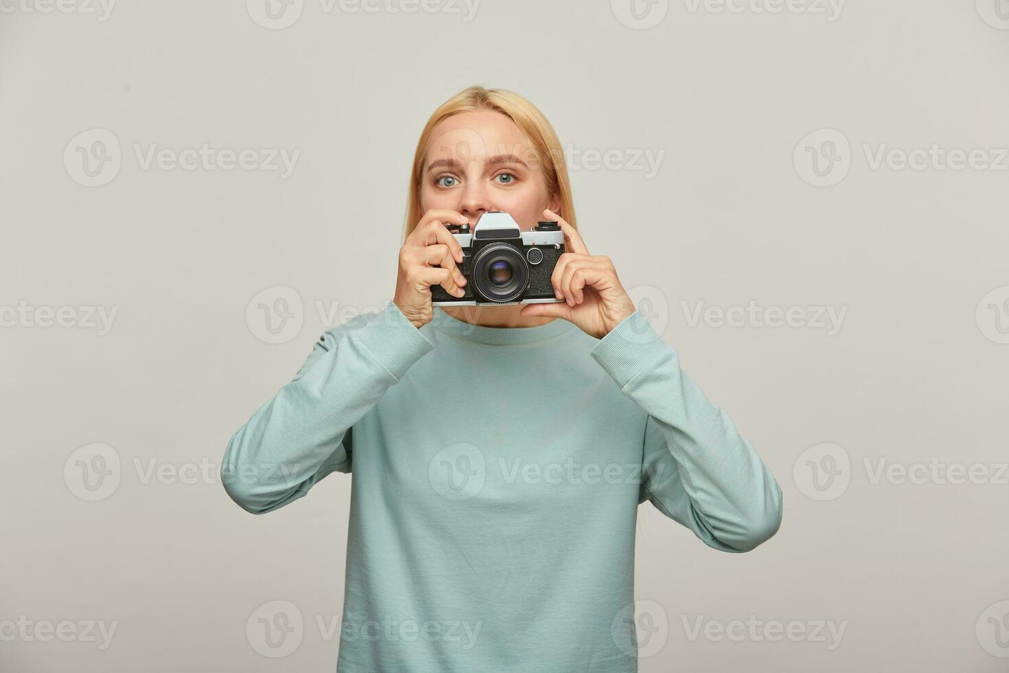 Young girl photographer looks out from behind the lens, holding a retro vintage photo camera, in the process of shooting, while working in studio, wearing blue casual sweatshirt, over grey background