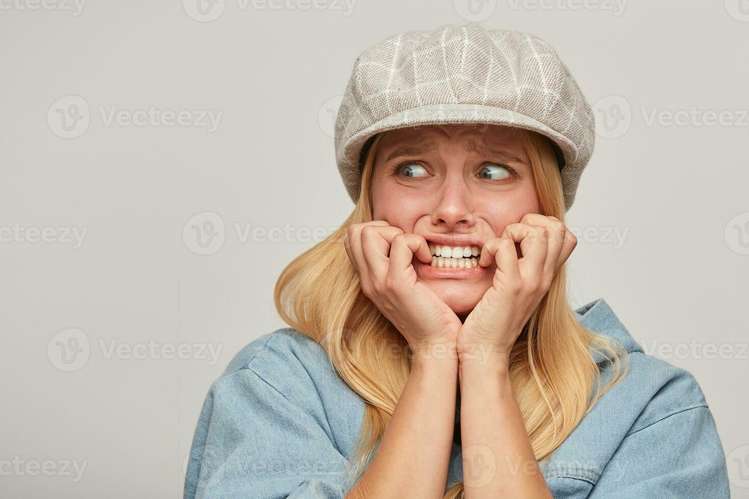 Scared blonde girl looks aside frightened afraid chattering teeth with fear,bites nails from fright, hands holding cheeks, on grey background photo